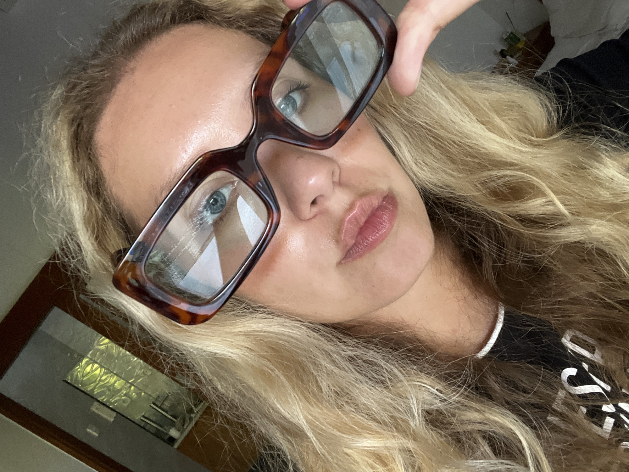 A close-up, high-angle, selfie-style photograph shows a young Caucasian woman with blonde, wavy hair. She is wearing large, tortoiseshell-patterned glasses and a black t-shirt with white text that is partially visible. Her light blue eyes are visible through the lenses of her glasses, and her lips are slightly parted. The background is blurred, but appears to be an indoor setting with a doorway and possibly a kitchen area reflected in a window. The lighting suggests it is daytime. The woman is located in Mengwi, Indonesia.