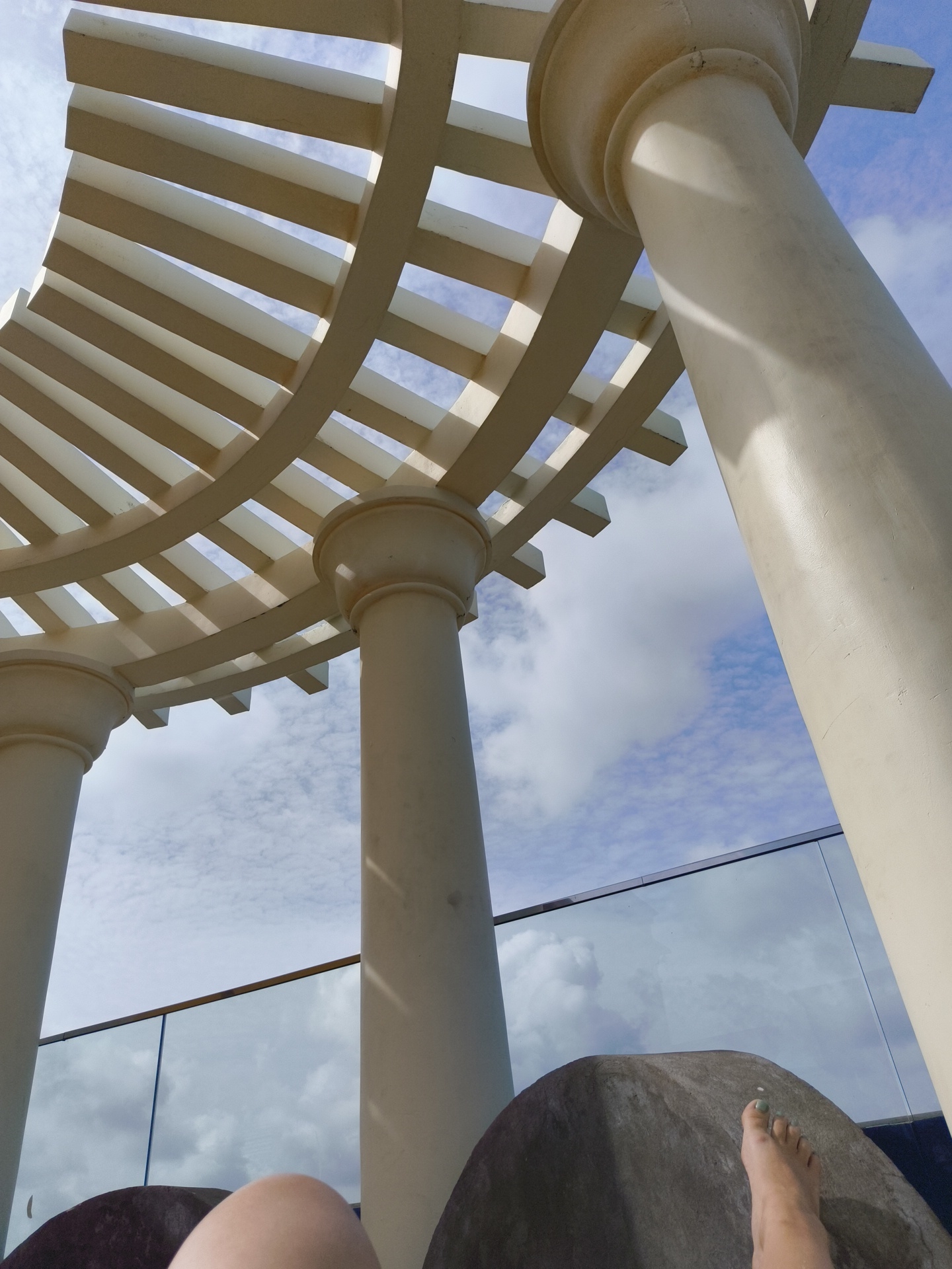 The image depicts a low-angle perspective of a beige pergola structure supported by two large columns. The pergola's slatted roof casts shadows on the sky, which is filled with white and grey clouds against a blue backdrop. In the foreground, a person's feet are visible, resting on a textured, dark grey surface, possibly a lounge chair or seating area. A glass railing is visible beyond the seating, suggesting an elevated location, such as a balcony or rooftop. The overall environment appears to be an outdoor setting, likely in a resort or public space, with a clear sky and architectural elements. The scene is likely in Phú Quốc, Vietnam, given the context of the query.