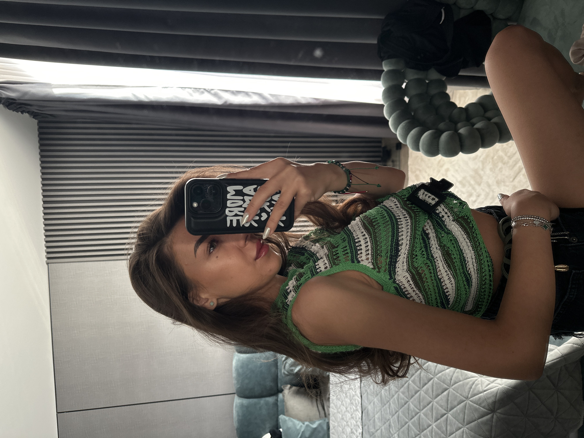 A young woman with long, wavy brown hair is taking a selfie in a room. She is wearing a sleeveless, crocheted green, white, and black top and dark shorts. She has on a silver bracelet on her left wrist. The background shows a bed with a quilted headboard, a textured wall with vertical slats, and a large gray curtain. A chair with a rounded, bubble-like design is visible on the right. The image was taken in Sofia, Bulgaria.