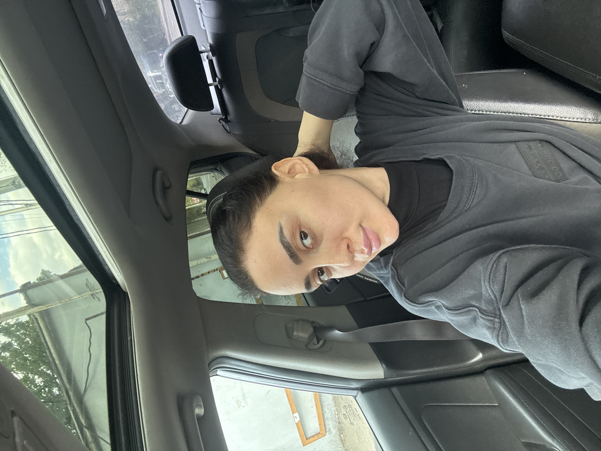 A person is sitting in the backseat of a car. The car appears to be parked near a building with a metallic exterior and a window showing foliage. The location could be Tbilisi, Georgia. The person is wearing a dark-colored sweatshirt or long-sleeved shirt. They are looking directly at the camera. The interior of the car is visible, including the seat, the headrest, and parts of the dashboard and door.