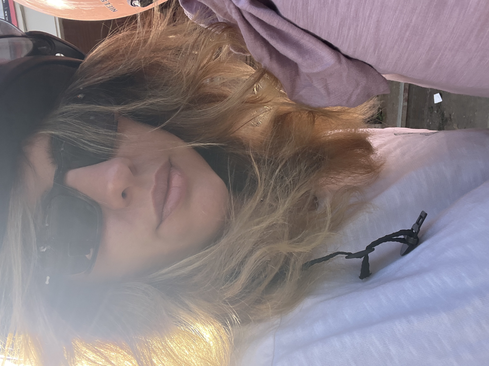 The image is a close-up, outdoor, selfie-style photograph taken in Kediri, Indonesia. The foreground is dominated by the face of a woman wearing sunglasses and a helmet. Her blonde hair is visible, catching the bright sunlight which creates a lens flare effect. To the right of the woman, another person is visible, wearing a pink shirt. The background appears to be a street scene with some greenery and a wall. The overall lighting suggests it is daytime, likely late afternoon due to the warm tones of the sunlight.