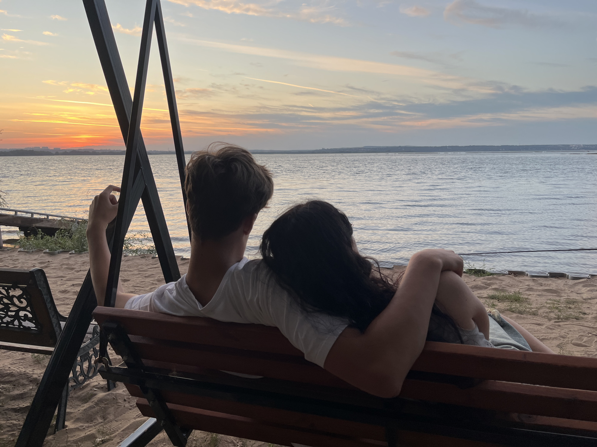 In Shumeika, Russia, a young couple is captured from behind, seated on a wooden swing set bench. They are facing a tranquil body of water, likely a lake or river, under a sky painted with the soft hues of sunset. The man, with light brown hair, wears a white t-shirt and rests his left arm on the back of the bench. The woman, with long dark hair, leans her head on his shoulder, her right arm draped across his chest and her left arm bent at the elbow. She wears a light-colored garment. The sand beneath the swing is lightly textured, with sparse patches of dry grass. In the distance, a faint shoreline with buildings is visible, silhouetted against the vibrant orange and pink sky. The water is calm, with a gentle ripple. The overall mood is one of peaceful intimacy and quiet contemplation of the natural beauty.