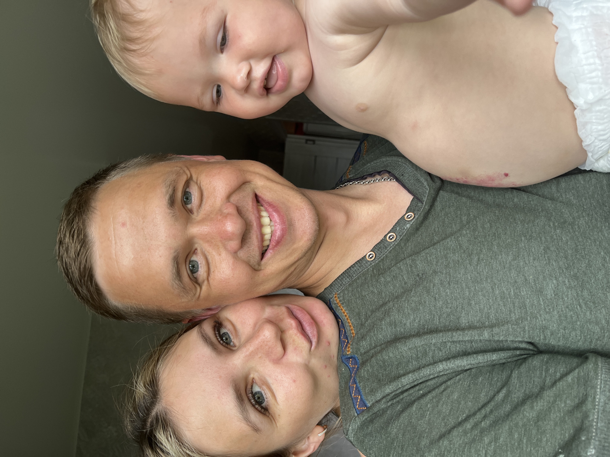 A close-up, informal portrait depicts a light-skinned man, woman, and baby in an indoor setting within the city of Chaikovsky, Russia.
The man, positioned centrally towards the right, has short, light brown hair, blue eyes, and a broad smile revealing his upper teeth. He wears a dark green, heathered Henley-style shirt with three visible dark buttons on the placket and a silver chain necklace visible below his throat. He is holding the baby on his right side.
To the left, a light-skinned woman leans her head on the man's right shoulder. She has light brown hair with blonde highlights, blue eyes, and visible freckles across her nose and cheeks. Several small, red blemishes are present on her left cheek and chin. She wears a small, silver hoop earring in her visible left ear and smiles softly towards the camera with slightly puckered lips. Her green garment features a blue, orange, and yellow geometric embroidered pattern along the visible shoulder and upper arm area.
The baby, held on the man's right, is light-skinned with fine blond hair, chubby cheeks, blue eyes, and a slightly open mouth with the tongue visible. The baby is shirtless, revealing a navel and a small, irregularly shaped reddish mark or lesion on the left side of the abdomen. A white, disposable diaper with a gathered elastic waistband is visible around the baby's lower abdomen. The baby's gaze is directed towards the right, away from the camera.
The background is a muted interior, with a light-colored, subtly textured wall visible on the left side and a darker, featureless area behind the man and baby, suggesting a shallow depth of field and soft indoor lighting.