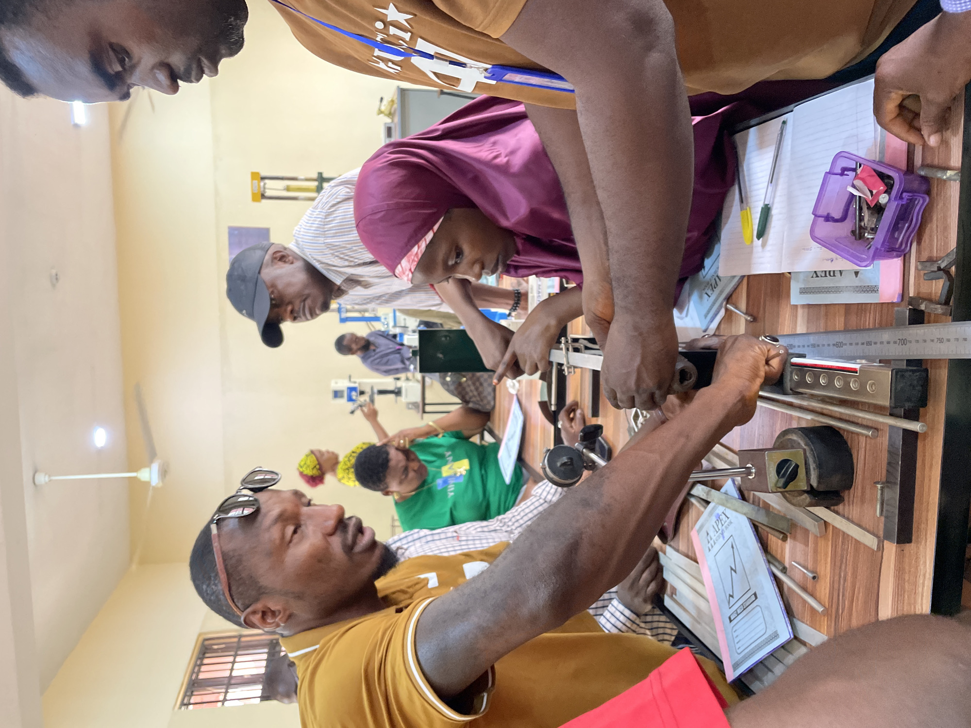 Afaka, Nigeria. A classroom setting. Individuals are gathered around a wooden table. A woman wearing a maroon hijab is observing or participating in a hands-on activity involving metal rods and a mechanical device. The group appears to be engaged in a practical learning exercise. Other individuals are visible in the background, including someone in a green shirt. The table has tools, notebooks, and writing instruments. The scene suggests an educational or training environment.