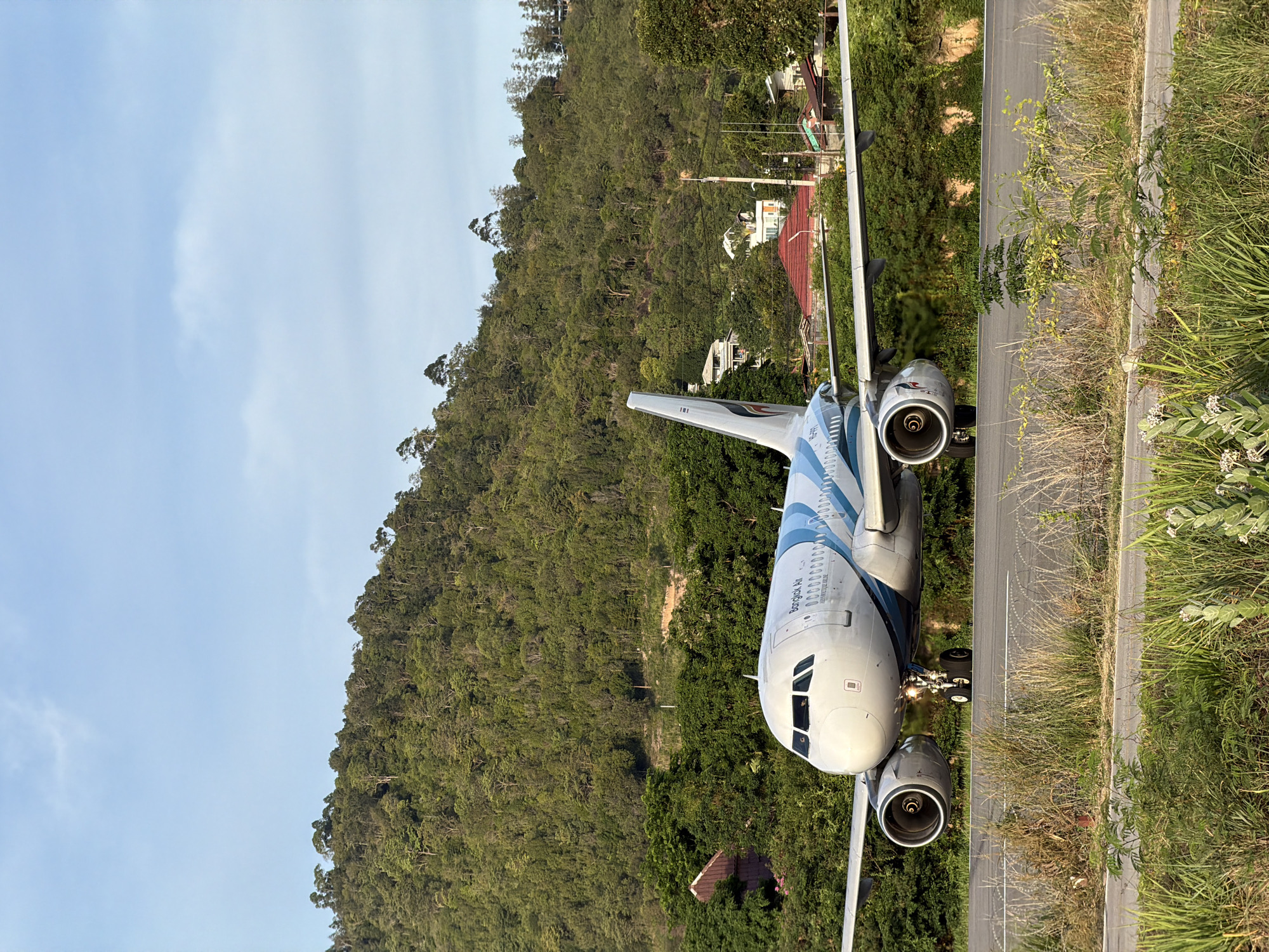 This media file shows a Bangkok Air airplane parked on a tarmac next to a tree-covered hill. The plane's nose is facing towards the camera, and its engines and wings are clearly visible. The plane is white with blue accents. The tarmac is paved and has white markings. The background is a dense green forest, with some houses visible among the trees. The sky is blue with some clouds. The image appears to have been taken during the day, likely in the morning or afternoon, given the lighting. The grass and vegetation along the edge of the tarmac are dry and tall, suggesting a tropical climate. The overall scene conveys a sense of quietness and stillness, with the plane appearing to be stationary. Visible text on the plane reads "Bangkok Air".