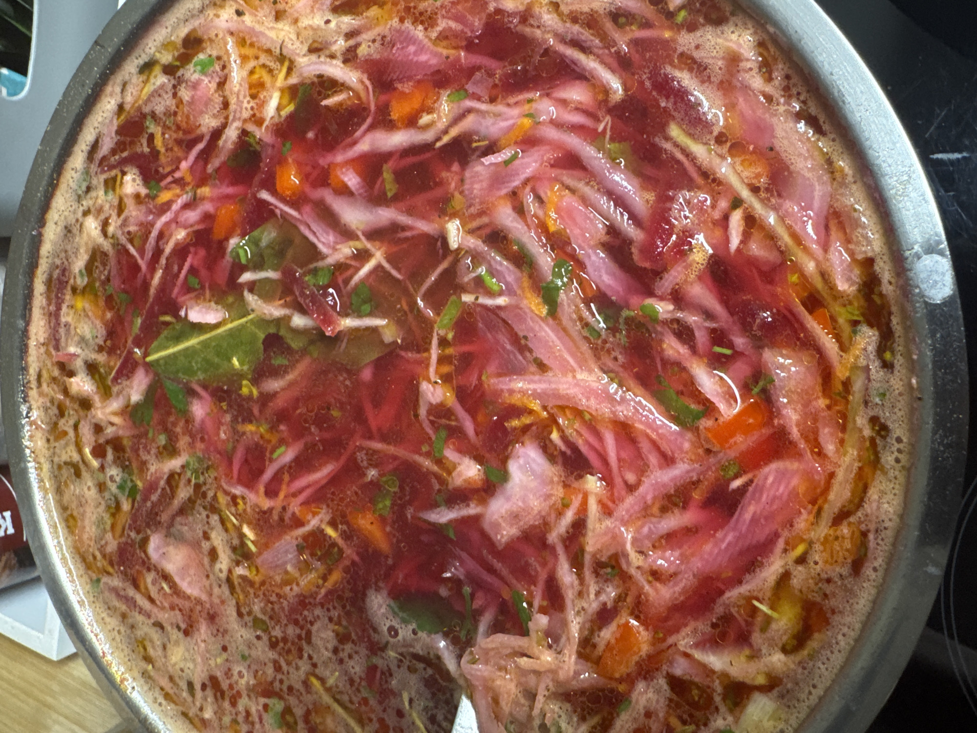 A close-up, overhead shot shows a stainless steel pot filled with a simmering, reddish-purple soup. The soup contains thinly sliced beets, shredded carrots, chopped onions, and green herbs. A bay leaf is also visible floating on the surface. The soup is bubbling and appears to be actively cooking on a black stovetop. The surrounding environment is out of focus but suggests a kitchen setting, possibly in a residential building in Katowice, Poland.