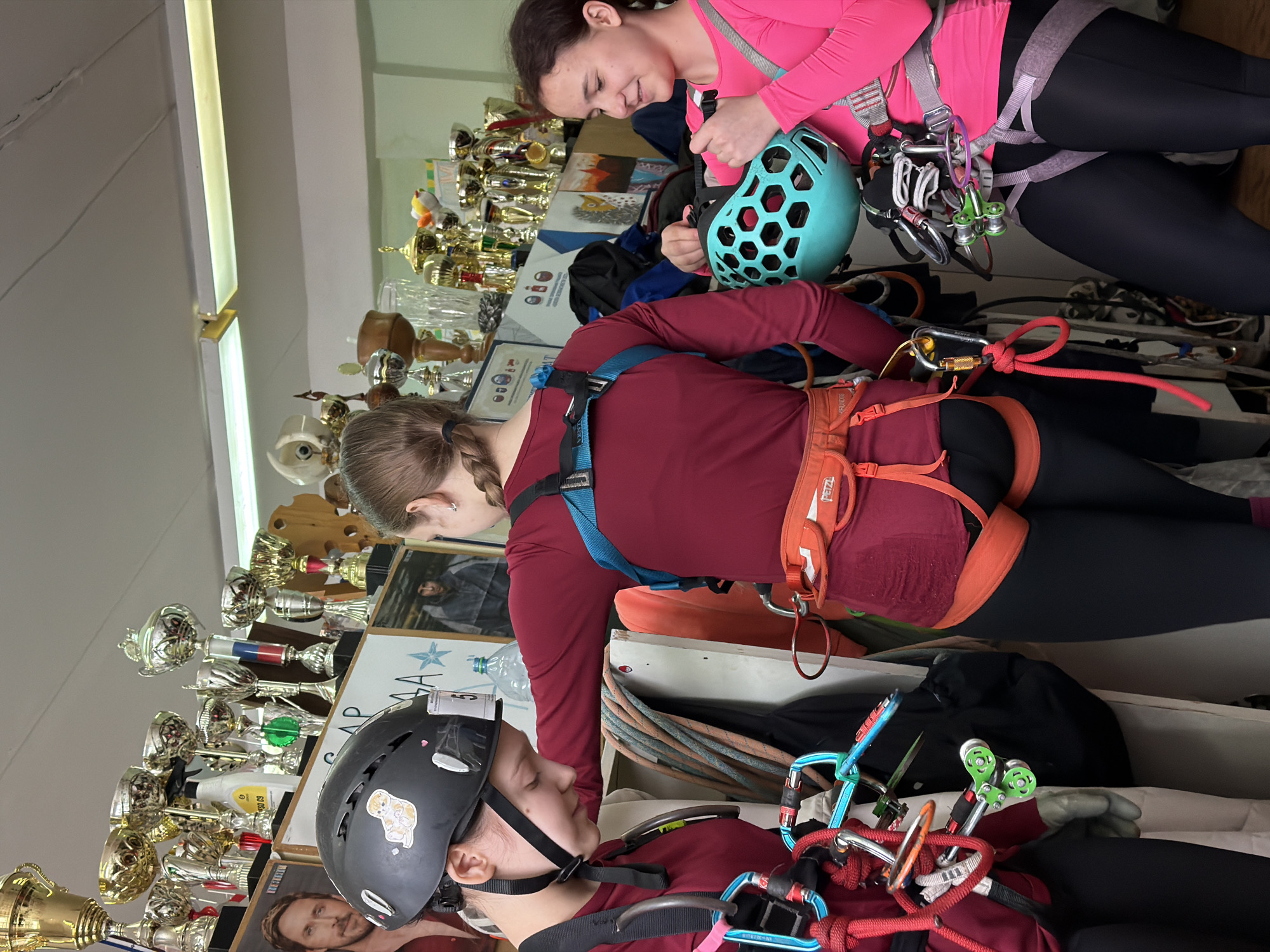 The image shows three people indoors. One person is wearing a pink long-sleeved shirt, black leggings, and a climbing harness. They are adjusting a blue climbing helmet. Another person is wearing a burgundy long-sleeved shirt, black leggings, and a climbing harness. They are looking down at a third person who is wearing a burgundy long-sleeved shirt, a black climbing helmet with stickers, and a climbing harness. Behind the people are shelves filled with trophies, photographs, and other climbing equipment. There is a fluorescent light fixture on the ceiling. The location is Berezniki, Russia.