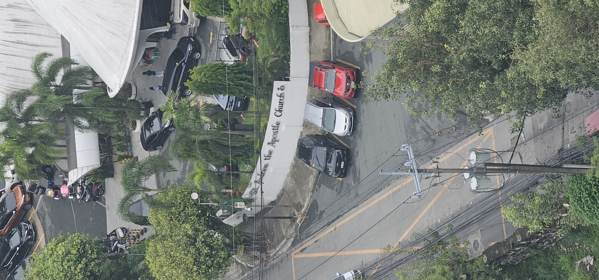 The image shows an aerial view of a street in Makati, Philippines, with a church sign that reads "St. Andrew the Apostle Church". There are several cars parked on the street and in a lot adjacent to the church. The scene includes trees, a utility pole with transformers, and a building with a large white roof. The weather appears to be overcast, and the street is wet, suggesting recent rain. In the foreground, a few people are visible walking near the church entrance.