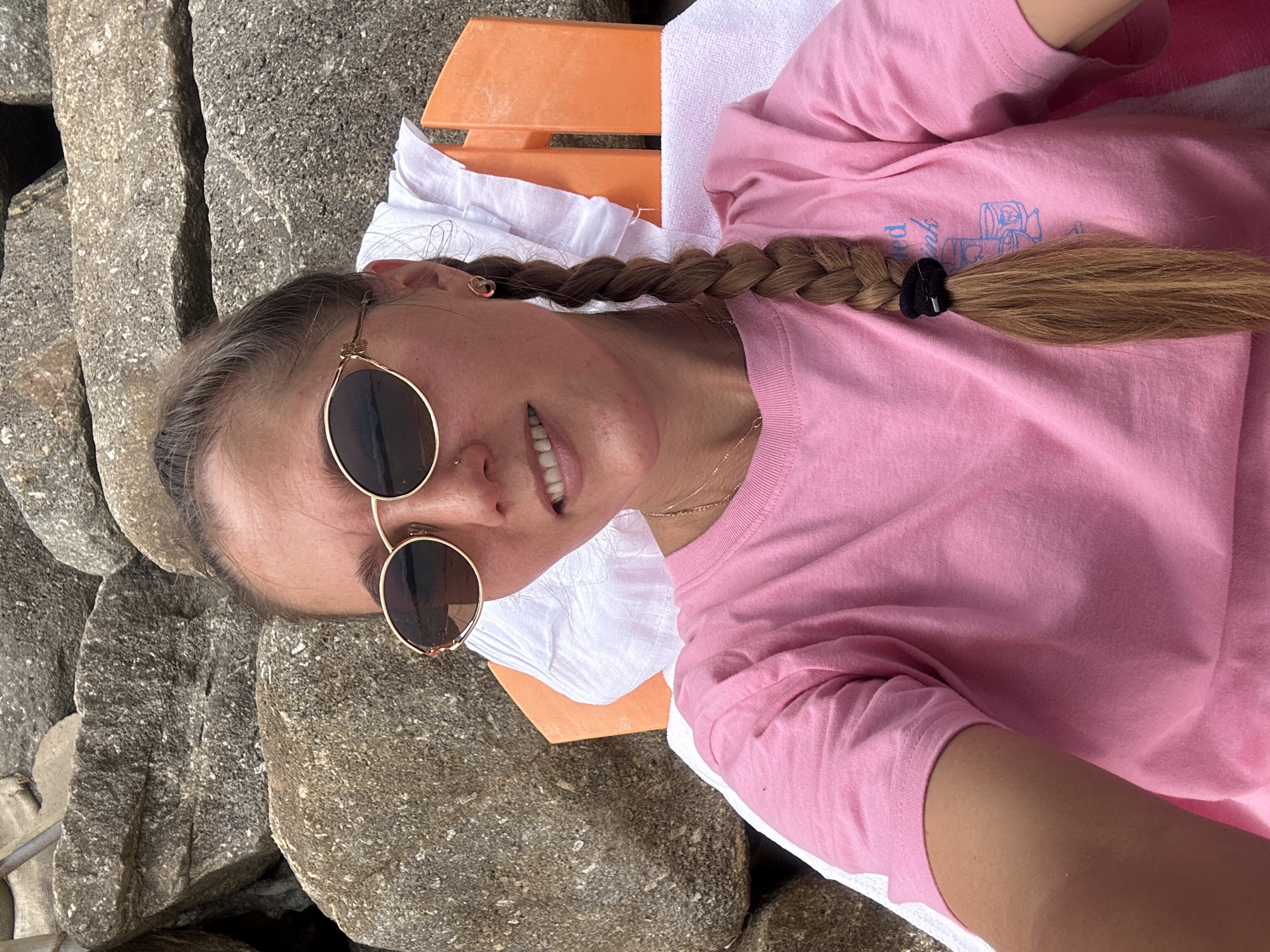 A young woman with blonde hair in a braid and sunglasses is sitting in a beach chair in front of a rock wall. She is wearing a pink t-shirt and a white towel is draped over the chair. The scene appears to be in Ko Samui District, Thailand.