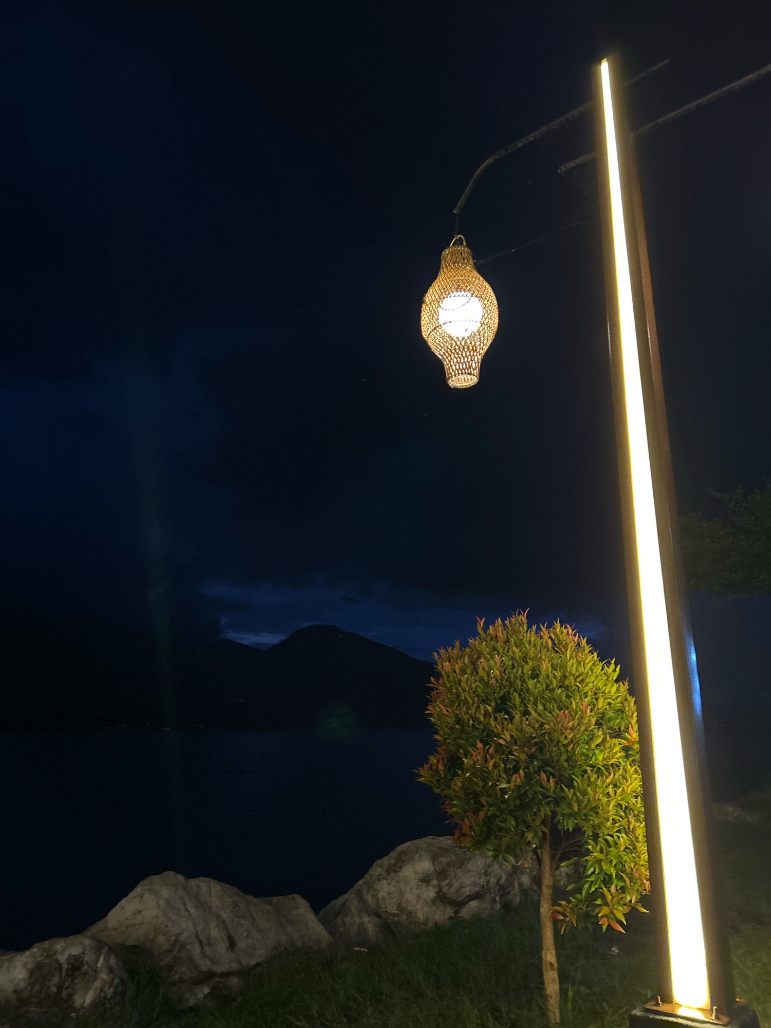 This image depicts a tranquil nighttime scene at the edge of a body of water, likely a lake, in Merodot, Indonesia. A decorative, woven lantern hangs from an arm extending from a tall, modern light post that emits a bright, vertical yellow glow. In the foreground, natural elements are visible: large, rough-textured grey rocks sit on a patch of green grass. A small, bushy green tree with reddish-tinged leaves stands to the right of the rocks, illuminated by the nearby light. In the background, a dark, expansive body of water stretches towards silhouetted mountains under a dark blue, cloud-filled sky. The overall ambiance is serene and atmospheric, suggesting an outdoor seating area or viewpoint at night. There are no people present in the image.