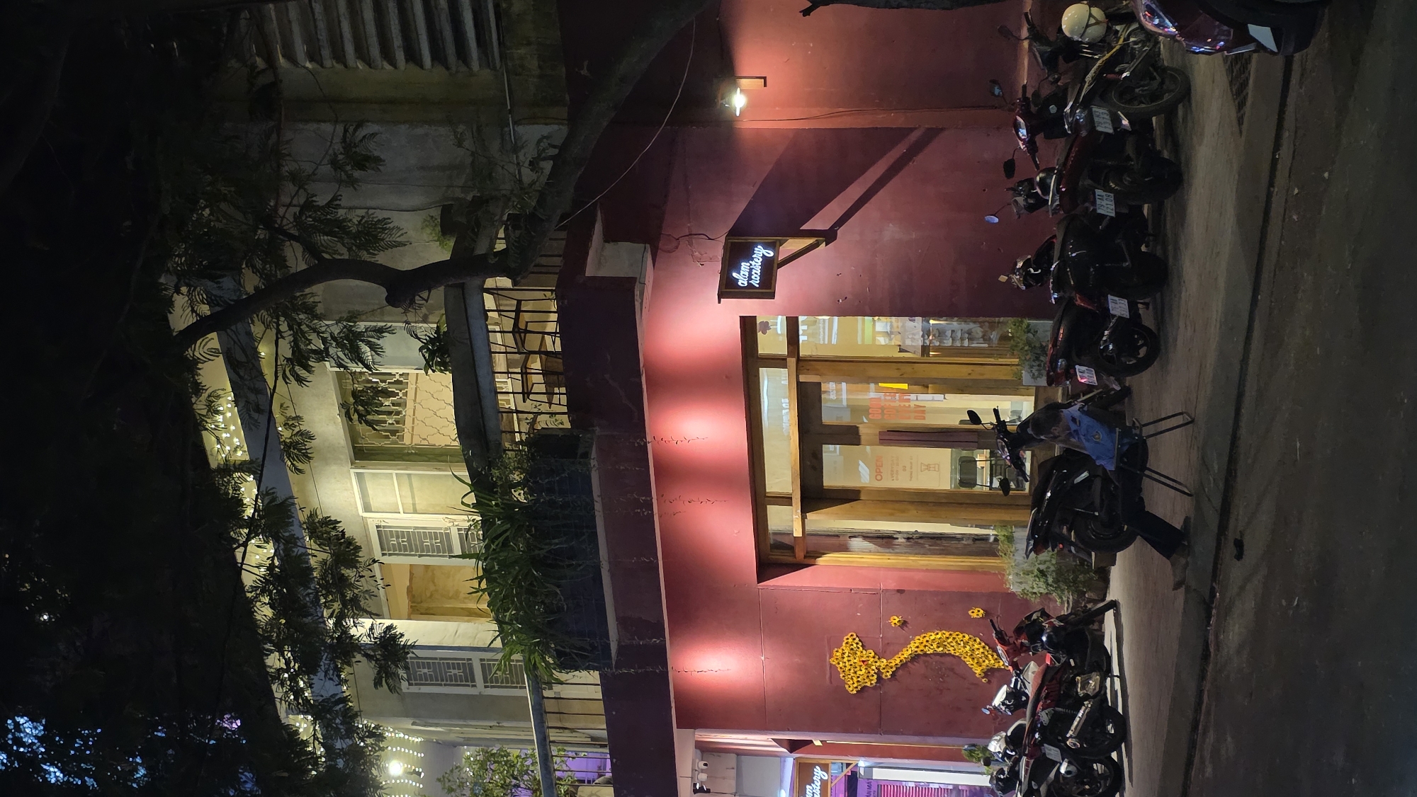 Captured at night in Nha Trang, Vietnam, the image shows a line of parked motorbikes on the right of the frame, positioned on a dark paved street. To the left of the motorbikes, a red building stands with an open-door revealing an interior with light. A lighted business sign that reads "alam roastery" hangs above the entrance. Below the entryway is a flower decoration. The building is next to a white-walled building with several windows. A tree with dense foliage is partially visible on the left.