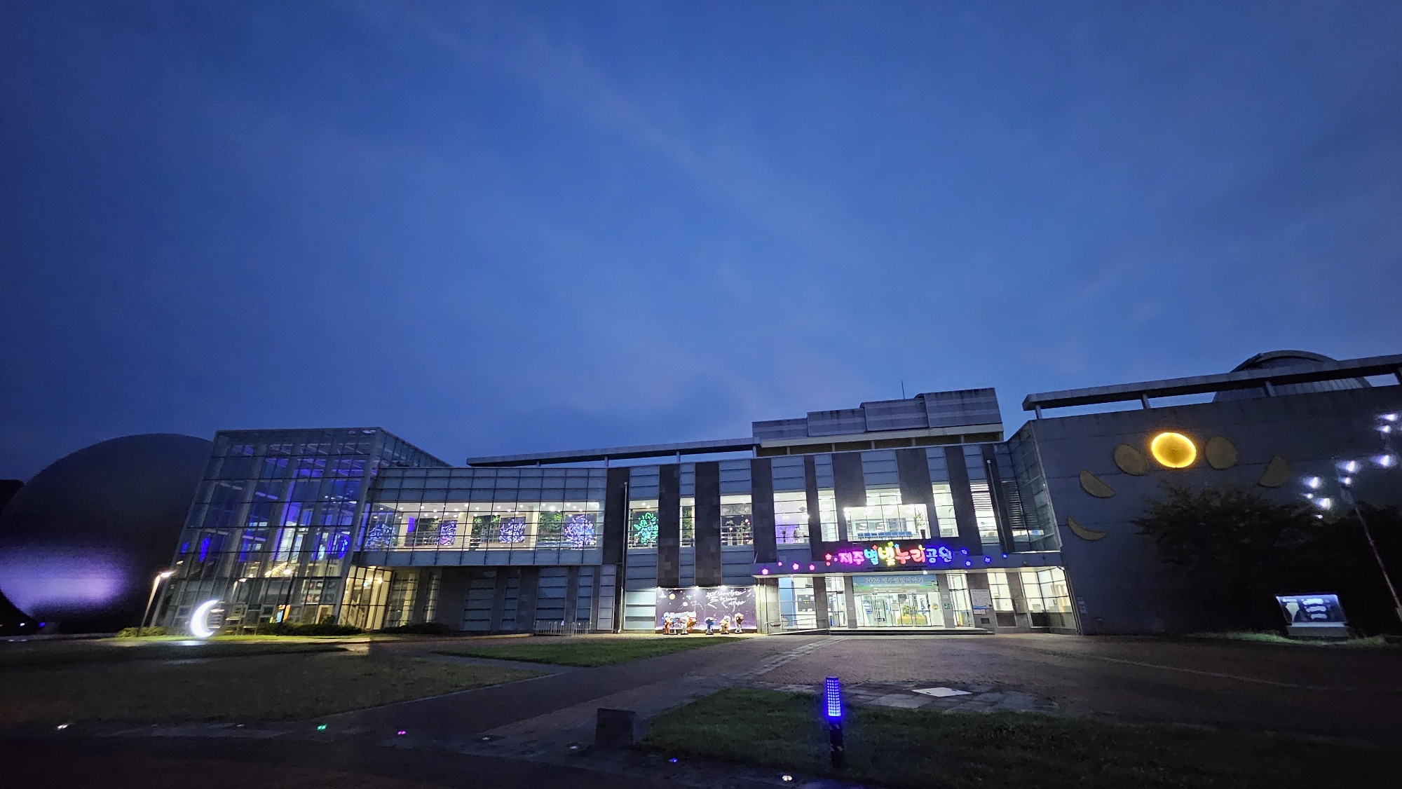 The image depicts the Jeju Science Center, located in Jeju City, South Korea, at dusk. The building features a modern architectural design with glass facades and geometric shapes. A large, domed structure, possibly a planetarium, is visible on the left side, illuminated with purple light. The main building is lit up from within, showcasing interior displays and a sign with Korean text. Decorative lights, including a crescent moon sculpture and small, colorful ground lights, enhance the exterior. The sky is a deep blue, characteristic of twilight. The surrounding area appears to be a park or landscaped grounds with grass and pathways.