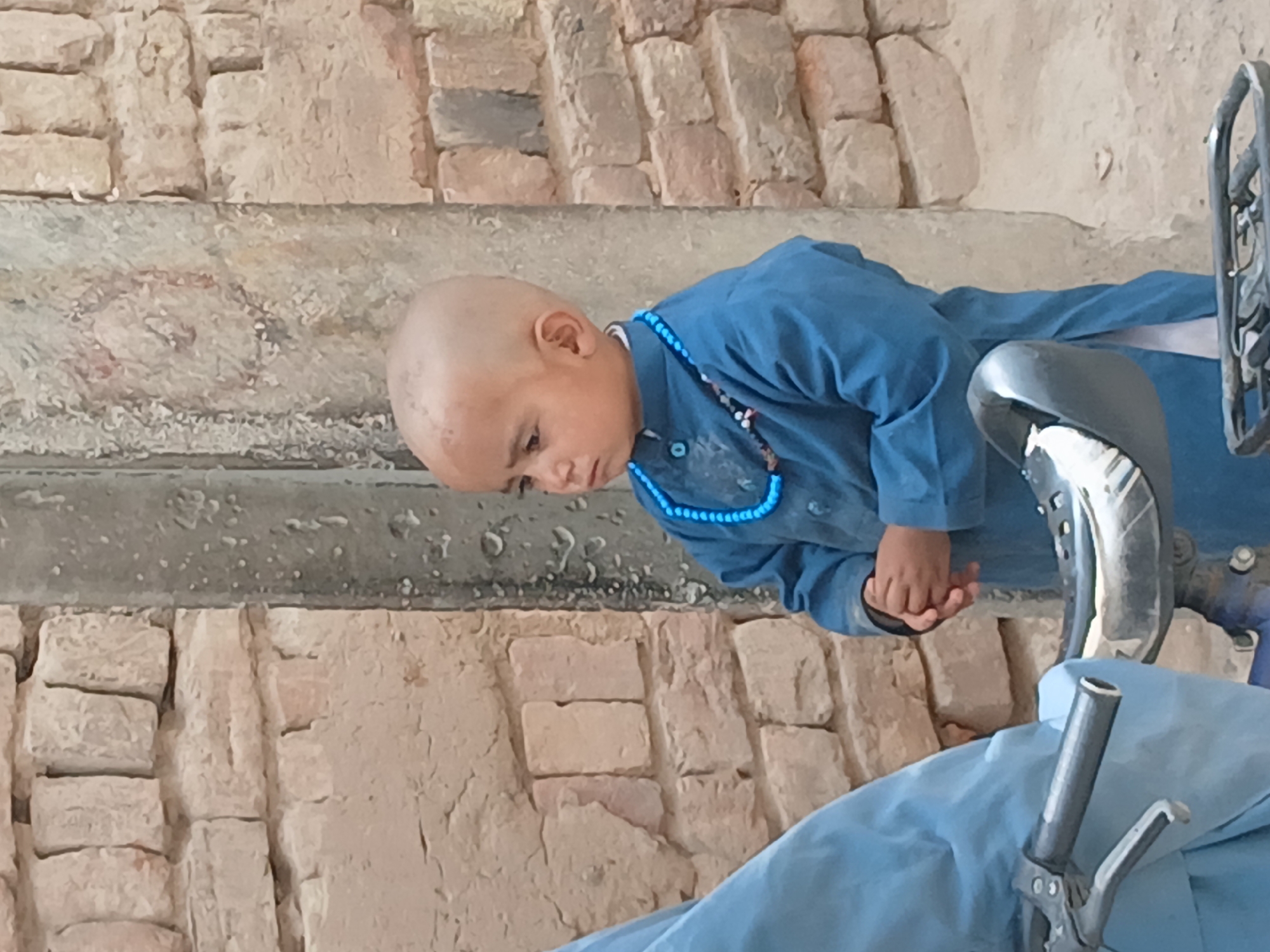 A young child with a shaved head, wearing a blue outfit and a blue beaded necklace, sits on a blue-covered surface. The child appears to be looking down at their hands. The background consists of a textured, light-colored brick wall, and part of a metallic object, possibly a bicycle or wheelchair frame. The location is Quetta City Tehsil, Pakistan.