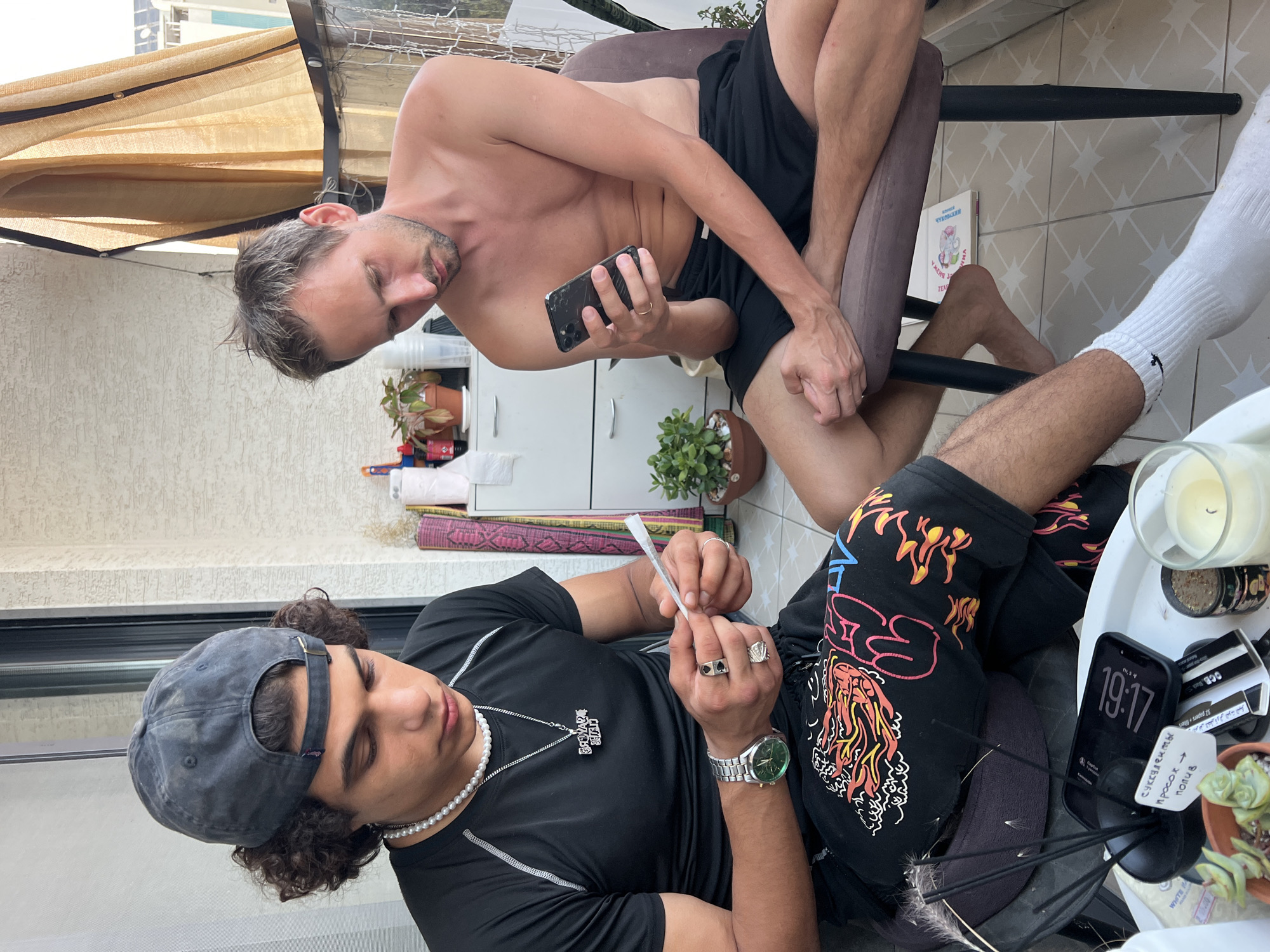 Two men are seated on a balcony in Kiryat Bialik, Israel, on a tiled floor with a textured wall behind them and a beige awning above. The man on the left, wearing a black t-shirt, a grey baseball cap, and black shorts with a colorful graphic print, is rolling a thin white object between his fingers. He has a pearl necklace and a silver pendant, a silver watch with a green face, and two rings on his hands. The man on the right, shirtless and wearing black shorts, is looking at his smartphone. He has a ring on his left hand. On a table in front of them are several items including a mobile phone, a lit candle, incense sticks in a holder, a small potted plant, and other miscellaneous objects.