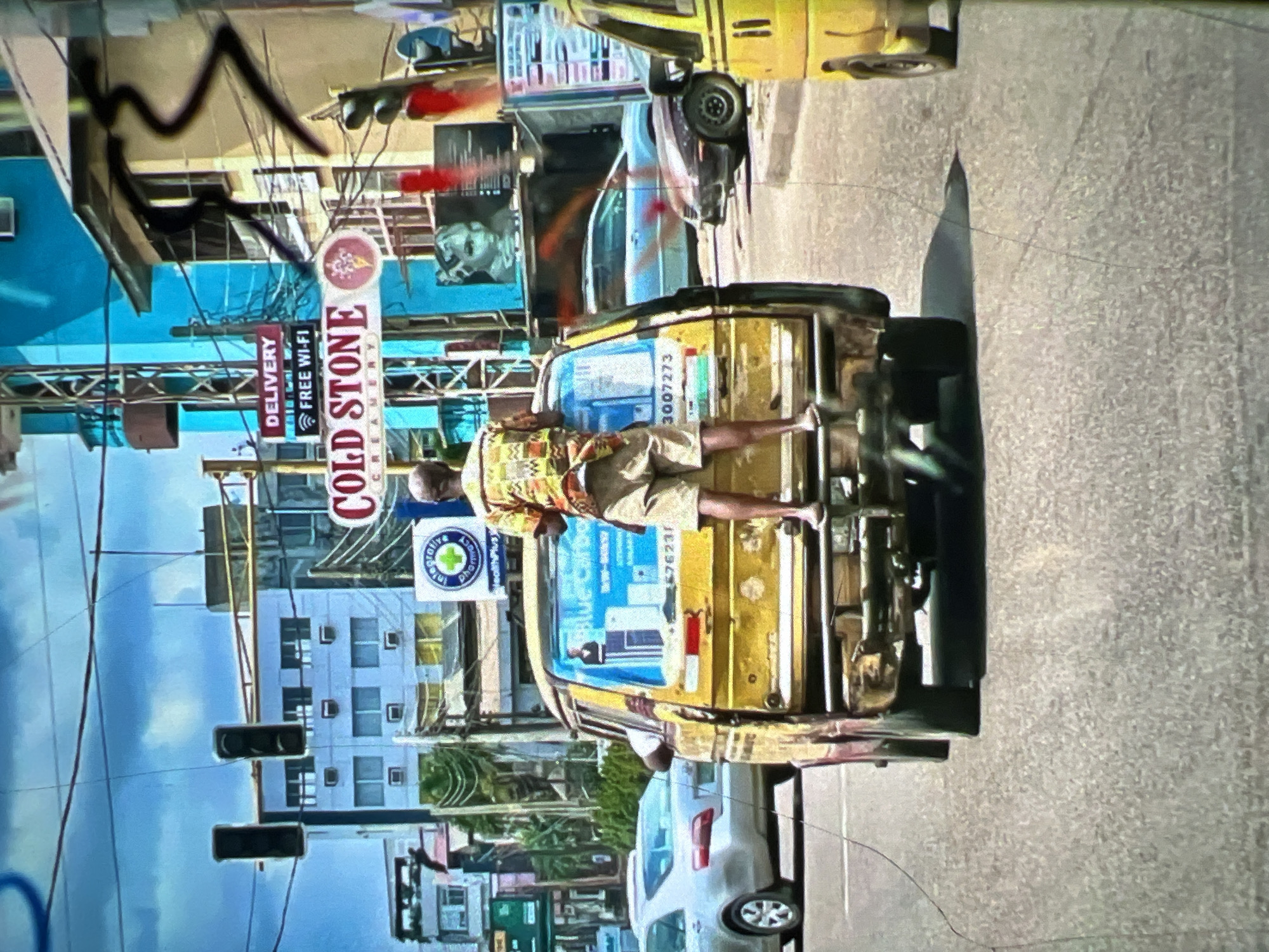 A man stands on the rear exterior of a yellow bus, his body angled towards the camera. He is wearing a patterned short-sleeved shirt and light-colored shorts. The bus is in motion, on a paved street with other vehicles visible. Buildings and signage line the street, including a sign for "COLD STONE CREAMERY" and another indicating "DELIVERY FREE WI-FI". The sky is blue with some clouds, and overhead wires are present. The overall scene depicts a busy urban environment in Lagos, Nigeria.