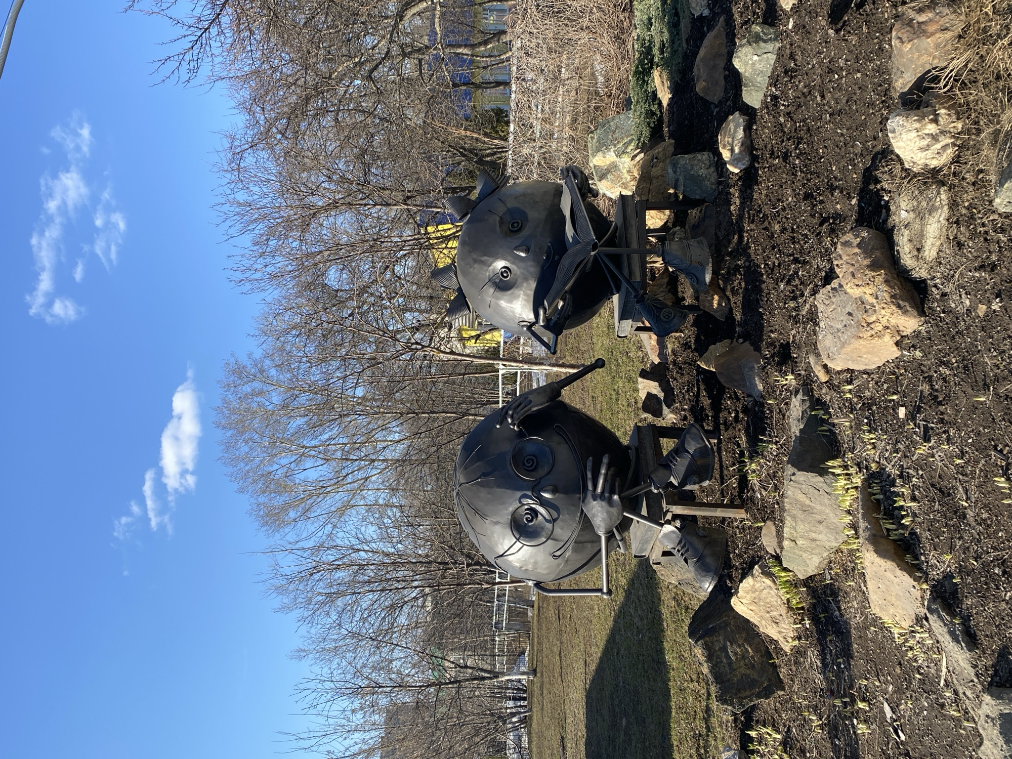 This image shows two metal sculptures in a public park in the city of Chaikovsky, Russia. The sculptures are whimsical figures with round heads, simple facial features (large eyes, small noses), and appear to be seated. They are positioned in a small landscaped area surrounded by rocks. The background includes bare trees, a blue sky with some clouds, and other park elements like fencing and buildings visible in the distance. The scene appears to be taken on a sunny day with clear weather.