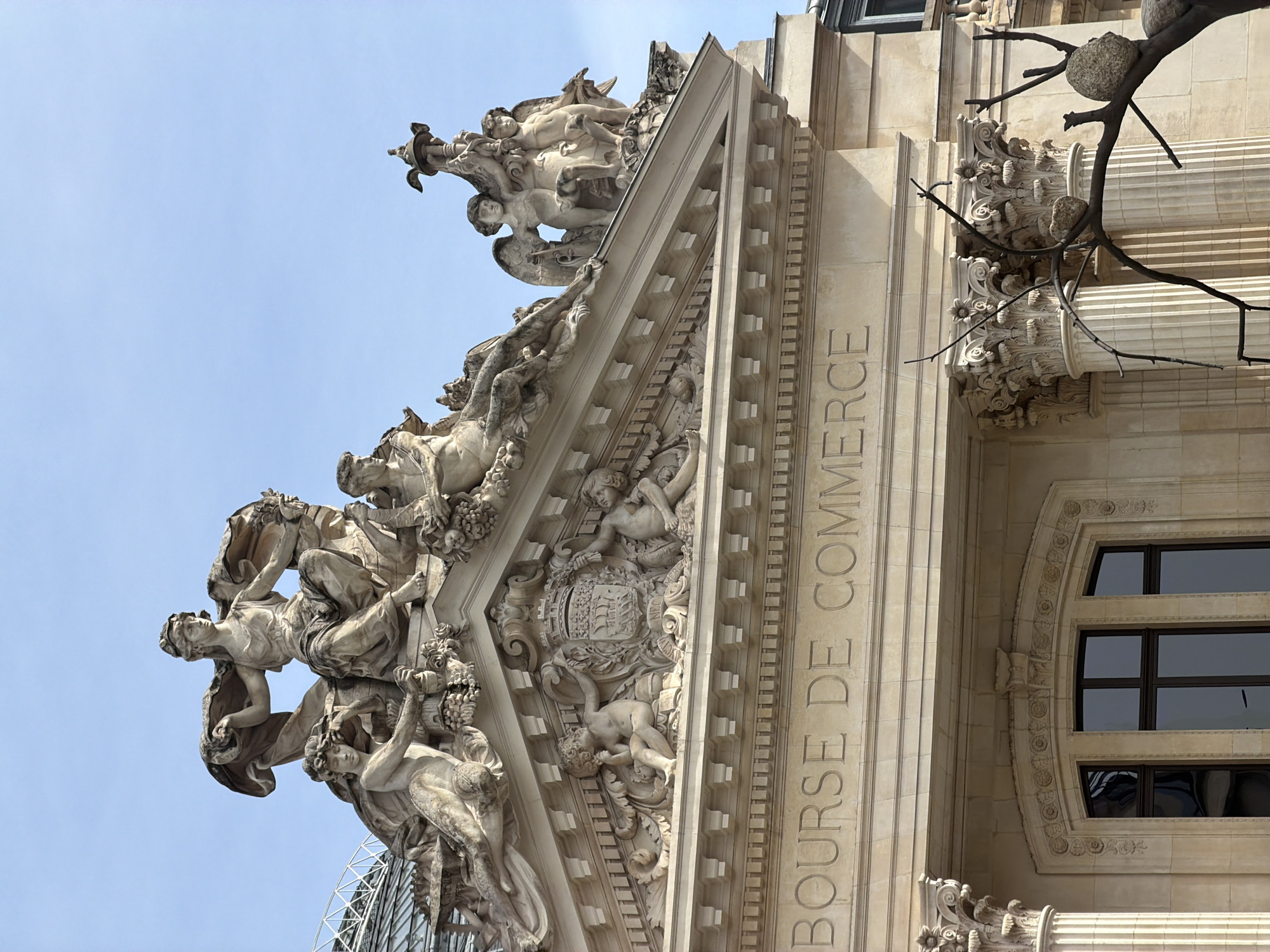 This image shows the upper corner of the Bourse de Commerce in Paris, France. The architecture is classical, featuring intricate stone carvings and sculptures. The words "BOURSE DE COMMERCE" are vertically inscribed along the building's facade. A group of statues adorns the building's top corner. A window is visible on the building, with decorative stone patterns. Part of the sky is visible in the background, along with a section of a tree with distinct balls attached to the branches. A modern steel-framed structure is visible below the stone structures.