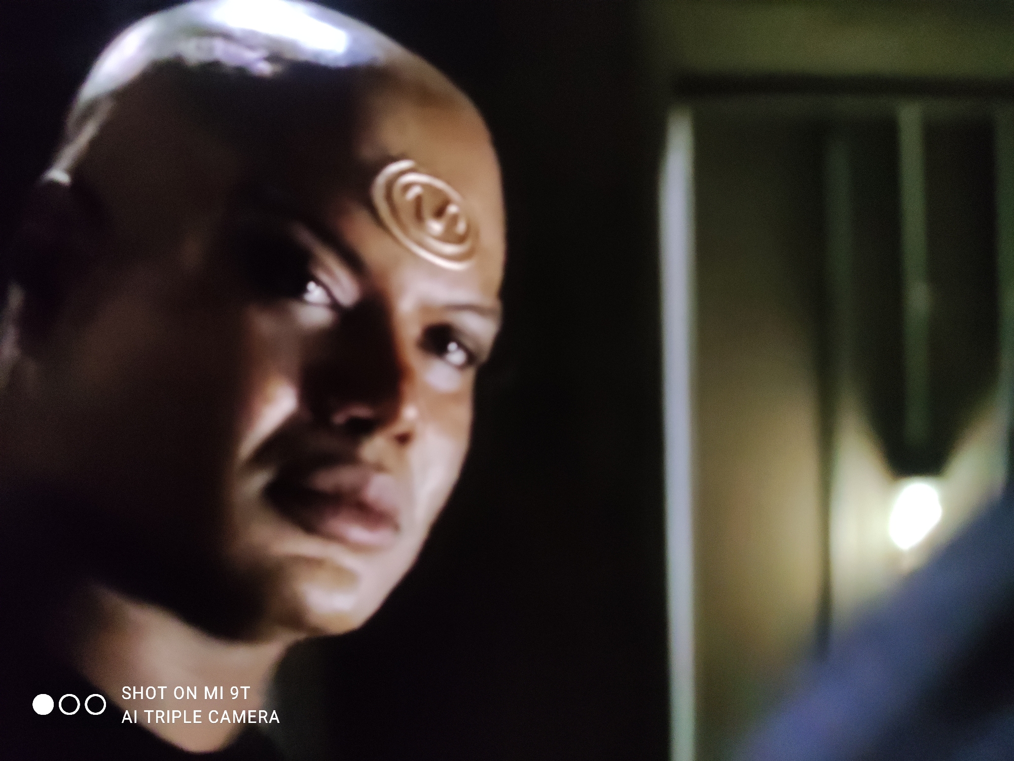 A close-up shot focuses on the left side of a bald person's face. The individual has dark skin and a prominent gold spiral symbol embedded in their forehead. Their left eye, dark and intense, looks directly at the camera. The right side of their face is in shadow. To the right of the person's head, a blurry, dimly lit background features vertical bars, suggesting a prison cell or a similar confined space. A bright, out-of-focus light source is visible in the far right background. The bottom left corner of the image contains a watermark that reads "SHOT ON MI 9T AI TRIPLE CAMERA". There is no information within the image to determine its location in Khazar Raion, Azerbaijan.