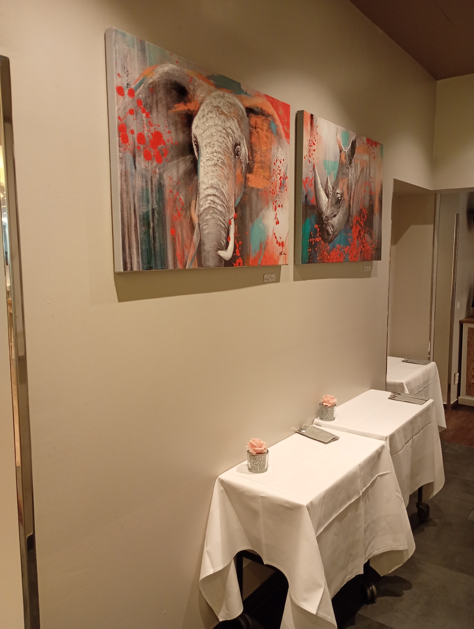 The image depicts an indoor scene, possibly a restaurant or dining area, featuring two large artworks of an elephant and a rhinoceros hanging on a light-colored wall. In the foreground, two tables are set with white tablecloths, cutlery, and small decorative pink flowers in glass vases. A full-length mirror is visible on the right side of the frame, reflecting another table that is also set. The floor appears to be dark tile. The lighting suggests an evening or dimly lit environment. The location is identified as the restaurant "Waldhaus am Schmelzteich" in Bad Sachsa, Germany.