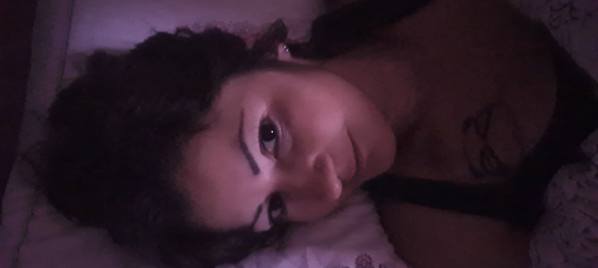 The image captures a young person, likely female, with dark, curly hair, lying down in a dimly lit indoor setting, presumably a bedroom. Their head is tilted, and they are looking directly at the camera with a calm and contemplative expression. The scene is cast in a soft, purple-pink ambient light, suggesting a low-light environment, possibly nighttime or a room lit by specific artificial sources. Dark streaks, resembling eyeliner or face paint, are visible beneath both eyes. On the person's upper right arm or shoulder, a dark, abstract or symbolic tattoo can be seen. They are resting on a light-colored pillow, with patterned bedding visible around them. The photograph offers an intimate glimpse into a quiet moment. No specific location cues are visible within the image, but the provided context places the scene in Lachish Regional Council, Israel. There is no visible text.