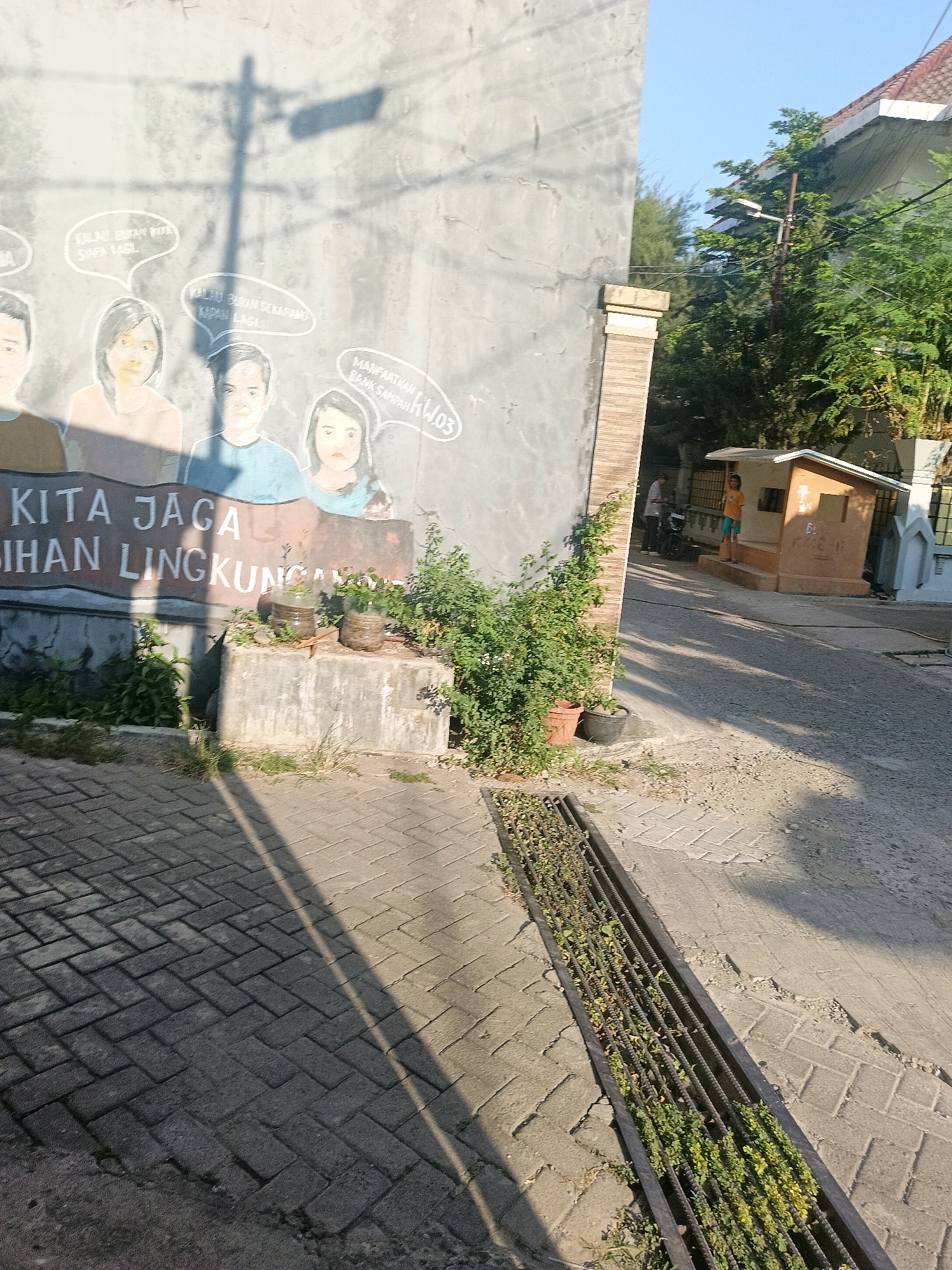 This is a photograph of a mural on a concrete wall depicting several figures with speech bubbles, alongside text reading "KITA JAGA" and "BIHAN LINGKUNGAN". The mural appears to be on the side of a building in Makassar, Indonesia, suggested by the distinct architectural style and vegetation visible in the background. In front of the mural, there is a low concrete platform with two small potted plants. Adjacent to the platform is a grey paved walkway, leading to a metal grate drain filled with green foliage. The scene is bathed in sunlight, casting shadows from a pole onto the mural and walkway. In the background, a small wooden structure with an orange roof is visible, along with trees and other buildings. A person in an orange shirt and blue shorts stands near the wooden structure, and another person and a motorcycle are partially visible further down the street.