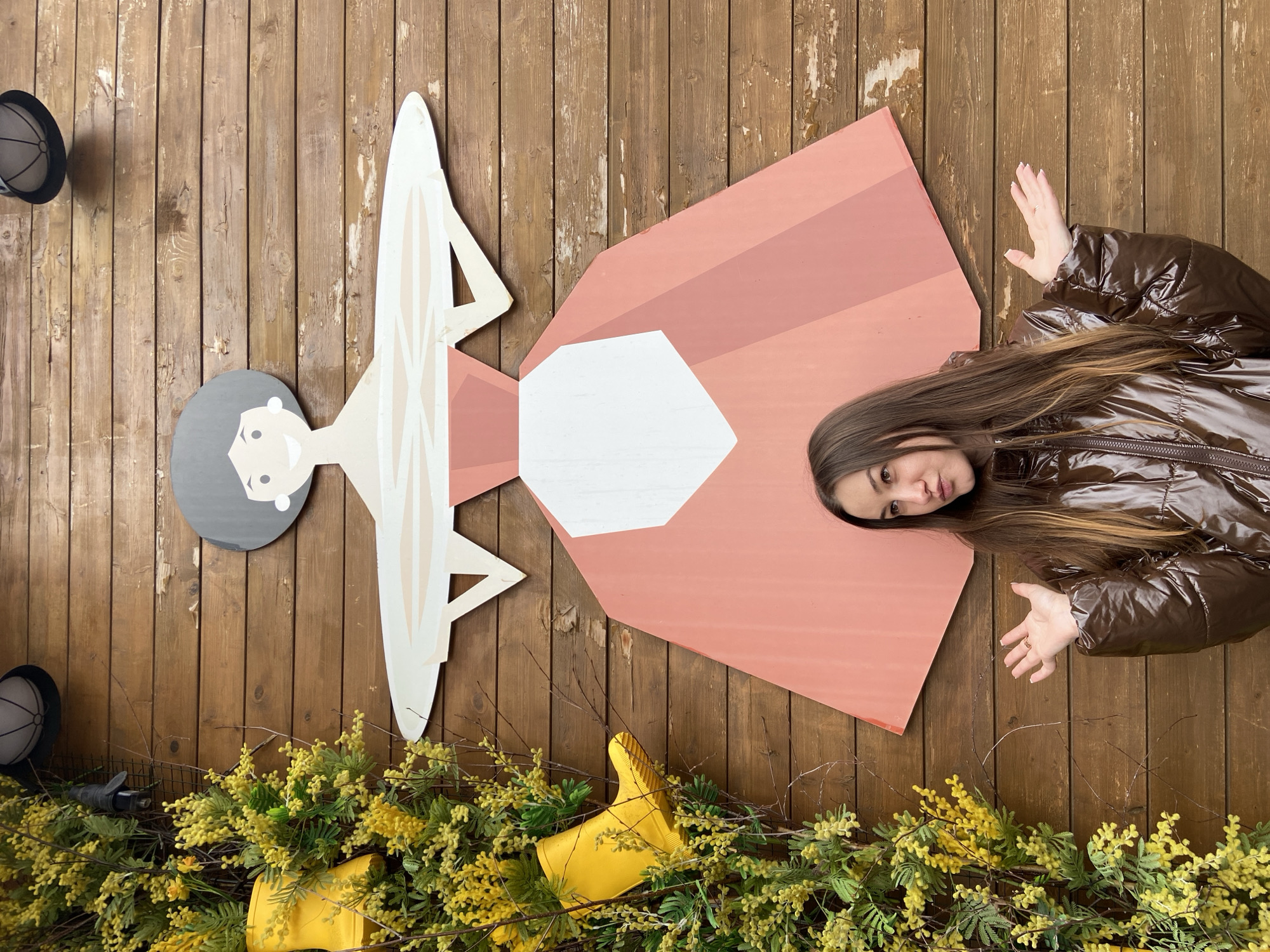 A girl in a shiny brown jacket poses with a life-size wooden cutout of a woman against a wood plank wall. The cutout is in the style of a simple, folk-art drawing of a woman with a grey head and white bodice and a rose-colored dress. The lower part of the image shows a border of yellow flowers and foliage, with a single yellow boot standing upright. The location is Izhevsk, Russia.