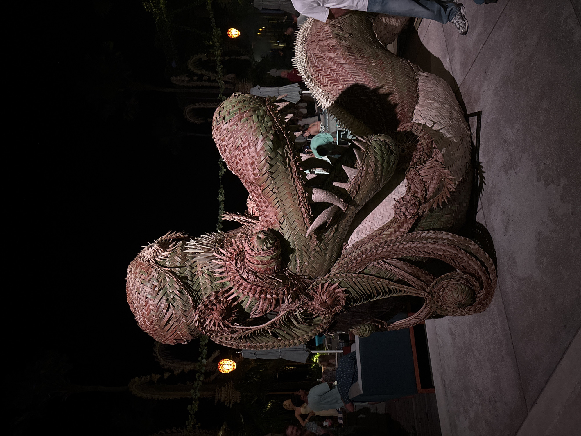 This night-time photograph from Kuta Selatan, Indonesia, showcases a massive, intricately woven sculpture dominating the scene. The sculpture, crafted from natural fibers like palm fronds, depicts a fierce, dragon-like creature with its mouth open, revealing rows of sharp, spiky teeth. Its body is composed of detailed, basket-like weaving in varying shades of reddish-brown and green, creating a textured, scaly appearance. The head and neck are particularly prominent, with numerous coiled and spiked details adding to its formidable presence.
In the mid-ground and background, several people are visible, gathered in what appears to be an outdoor or semi-outdoor venue, such as a restaurant or event space. They are casually dressed, some seated, and appear to be socializing. The setting is illuminated by artificial lights, including a warm, glowing lantern hanging in the background, which casts soft light on the surrounding area. The ground beneath the sculpture and people is a light-colored, paved surface, showing shadows cast by the figures and the sculpture. The dark, unlit background emphasizes the illuminated sculpture and the gathering. The overall impression is a lively, relaxed evening atmosphere, centered around the impressive and unique piece of natural fiber art. No visible text is present in the image.