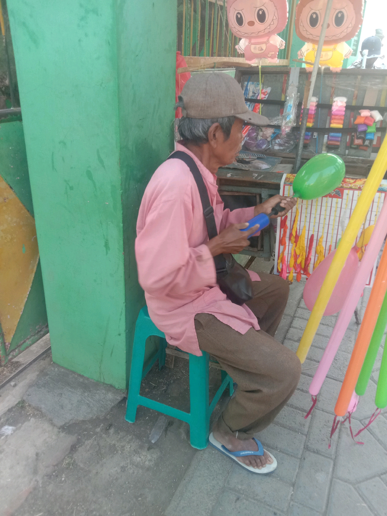 In Selotambak, Indonesia, a man sits on a green stool, wearing a pink long-sleeved shirt, brown pants, a brown cap, and blue and white flip-flops. He is holding a blue balloon pump and inflating a green balloon. The man is positioned in front of a stall that sells balloons and other toys. In the background, there are several colorful balloons, including two large character balloons, one pink and one yellow, and stacks of smaller balloons. The stall also displays various other items in plastic packaging. The man's bag is slung across his chest. The ground appears to be paved with concrete and brick. A green wall is visible to the left of the man, with a portion of a yellow and green patterned surface. Several long, brightly colored balloons (yellow, pink, orange, and green) are attached to sticks and stand upright next to the man.