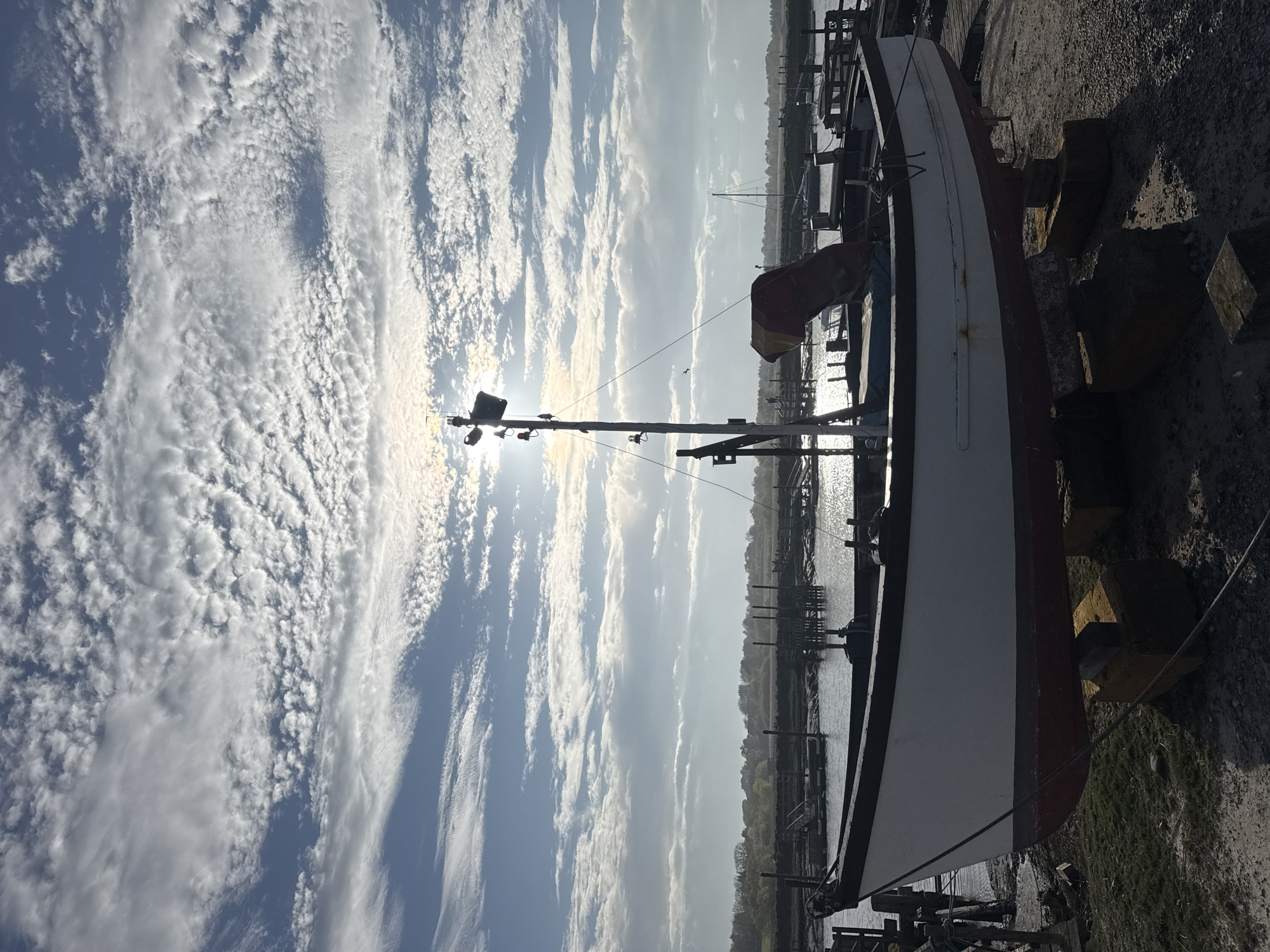The image shows a boatyard in East Suffolk, United Kingdom. A medium-sized, dry-docked vessel is the primary subject. It's painted white and red and sits on supports. To the left, the sky is filled with altocumulus clouds. In the background, a river or estuary reflects sunlight, and a line of trees is visible. The foreshore is gravelly. There are no people in the immediate view.