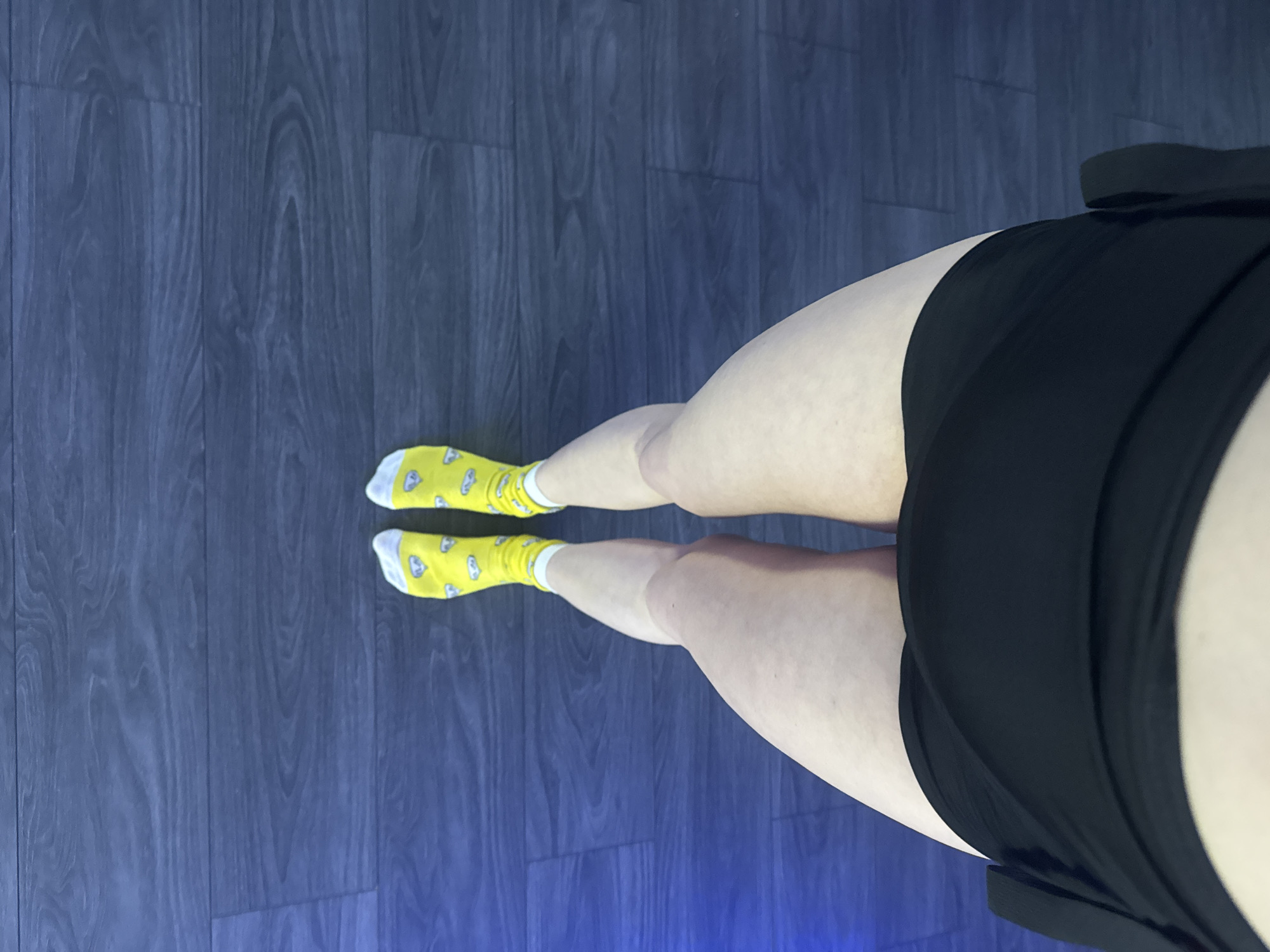 A high-angle shot shows a person's legs from the knees down, adorned with yellow socks patterned with white cartoon faces. The individual also wears black shorts. They stand on a floor composed of dark gray planks that appear to be made of wood or a similar material, oriented vertically in the image. The lighting in the area seems artificial, with a slight blue tint, particularly noticeable at the bottom of the frame. No other elements such as furniture or people are visible in the scene. The location and city are unknown.