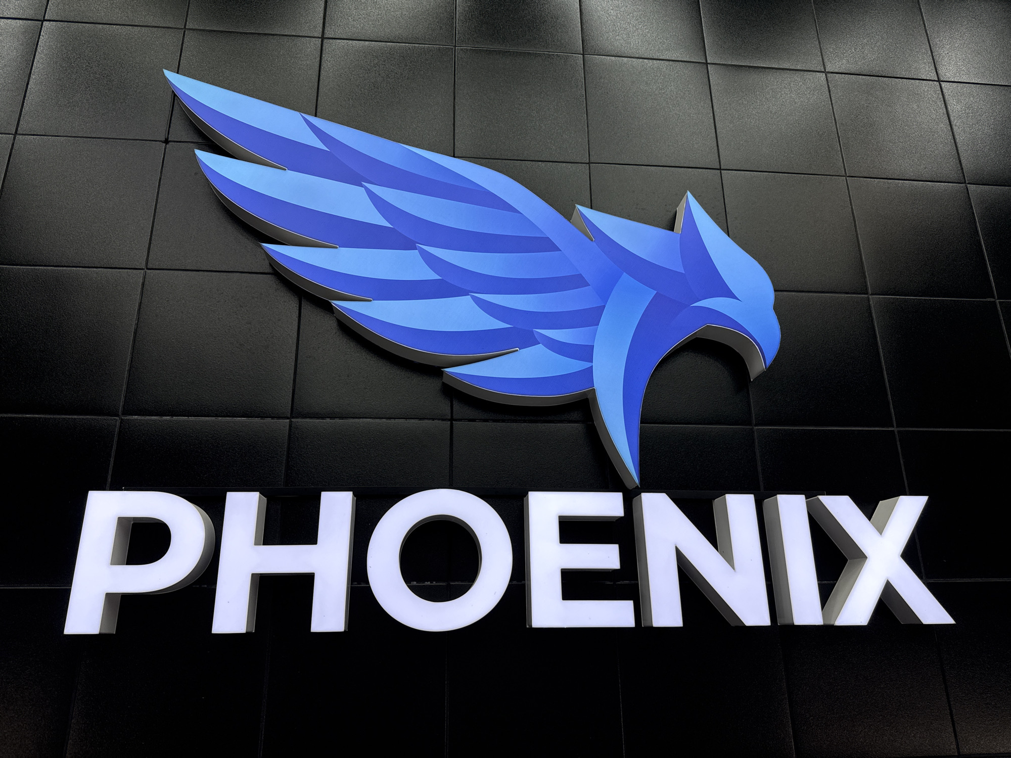 The image shows a wall-mounted sign that reads "PHOENIX" in large, illuminated white block letters. Above the text is a stylized emblem of a phoenix, rendered in shades of blue. The background is a black, tiled wall. The precise location in Saint Petersburg, Russia, cannot be determined from the image.
