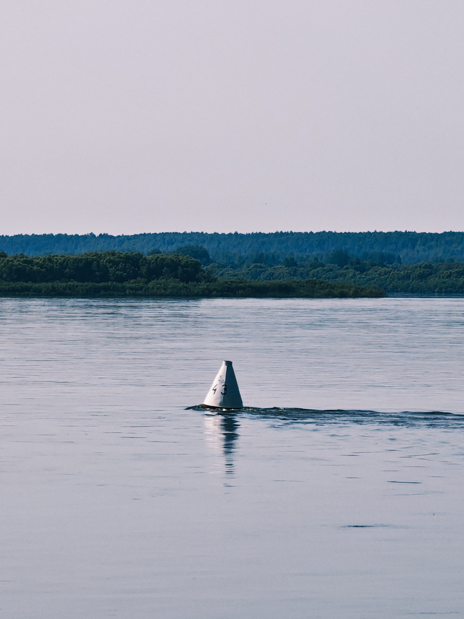 A conical white buoy with the number "43" in black script is partially submerged in calm water, casting a reflection. The buoy is positioned slightly off-center to the right of the frame. A faint wake trails behind the buoy, indicating movement. The water is a muted blue-gray, reflecting the pale sky. In the background, a dense, dark green treeline stretches horizontally across the horizon, forming the edge of a forested area. The sky above is a gradient of pale blue and white, with no visible clouds. The scene appears to be a tranquil outdoor environment. This image was taken in Novonazimovo, Russia.