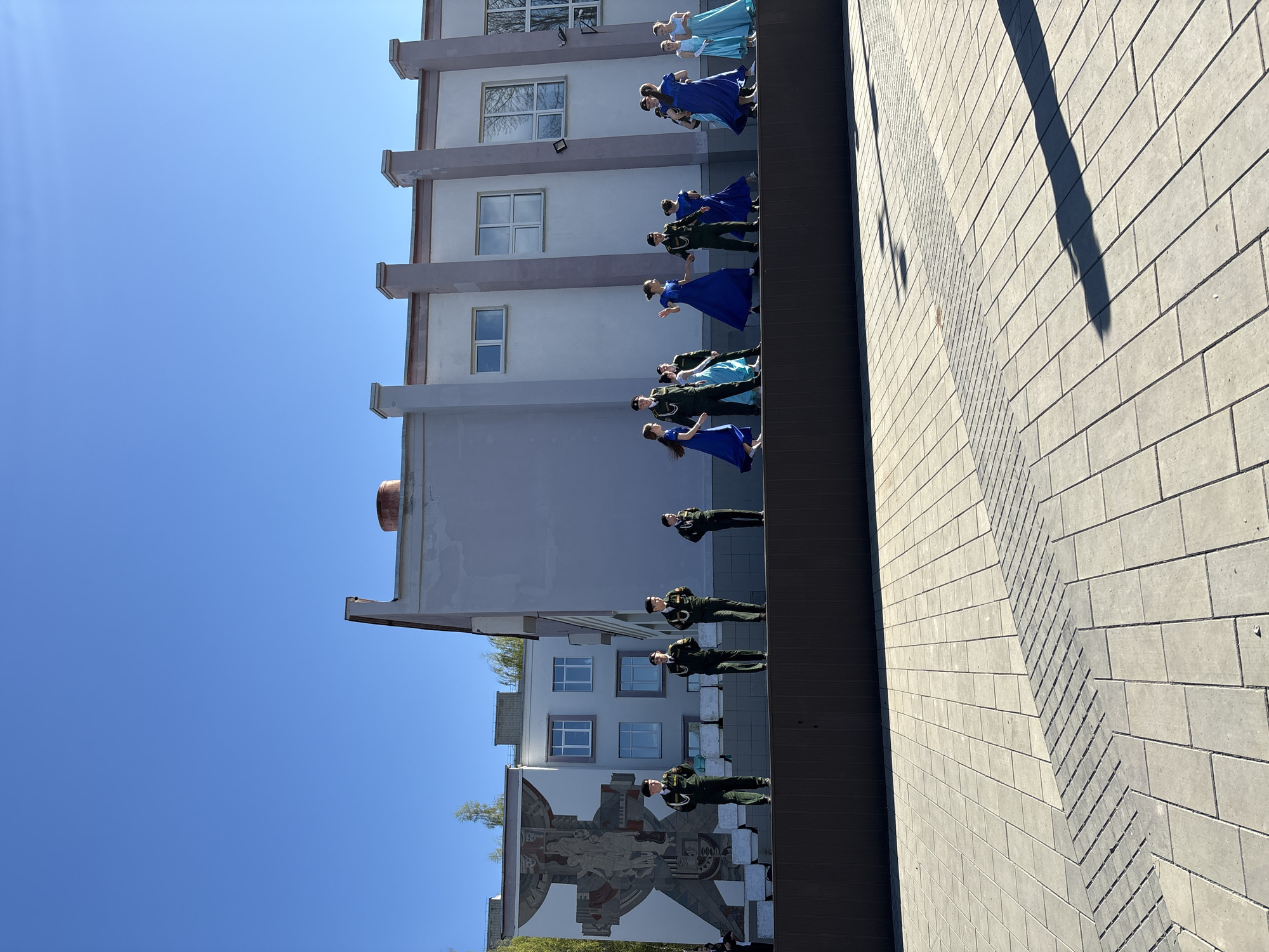 The image shows a row of people standing on a ledge in front of a building. Some of the people are wearing military uniforms, while others are wearing formal dresses, seemingly rehearsing or posing for photos. The building behind them is multi-story with windows and architectural details such as columns. A mural is visible on the lower part of the building. The location is identifiable as Chaikovsky, Russia based on the specific architectural style and mural details, which are characteristic of buildings found in the city. The ground in front of the building is paved with rectangular blocks. The sky is clear and blue.