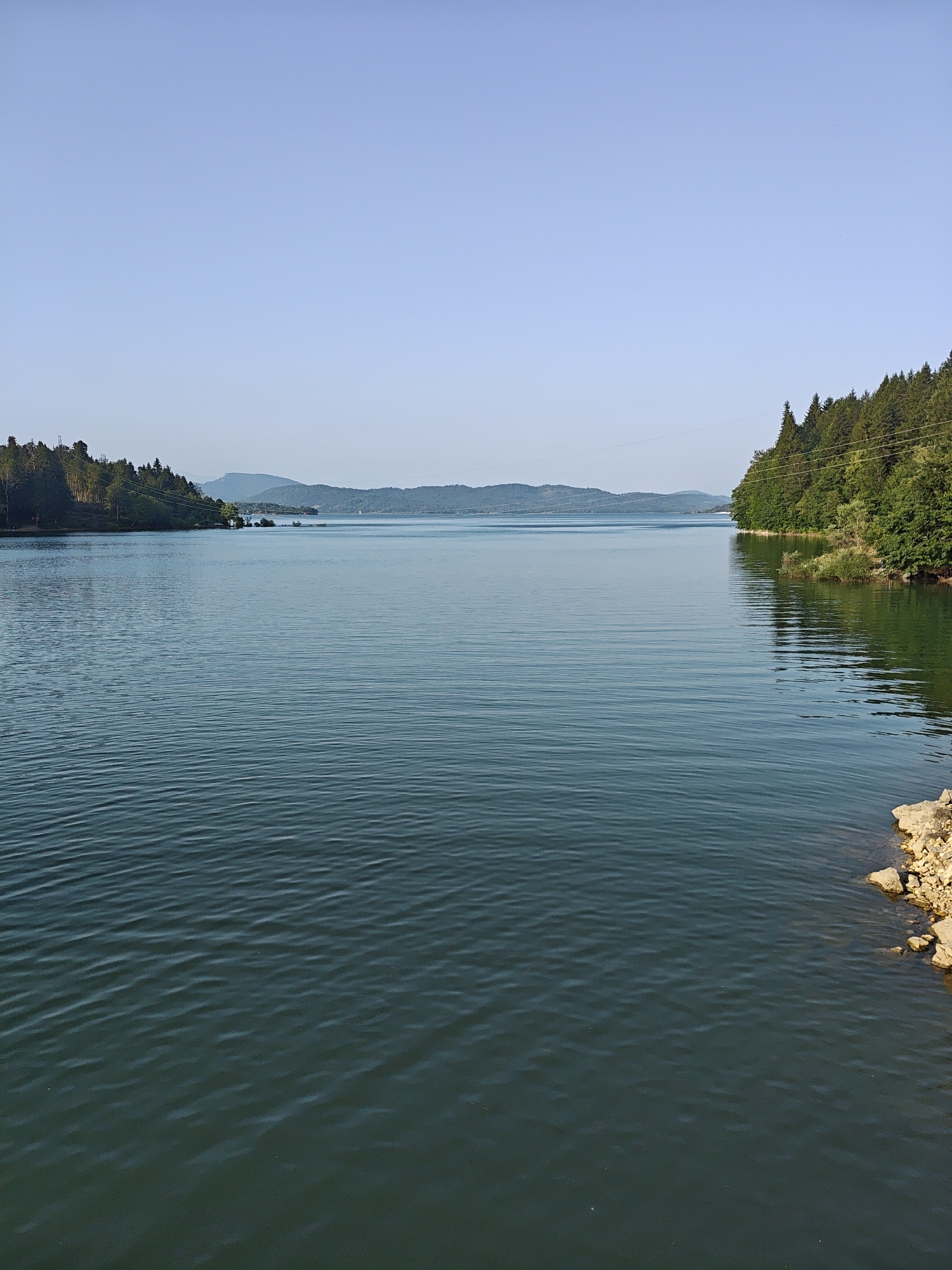 This is a wide, eye-level shot of a large, calm body of water, likely a reservoir, extending into the distance. The water is a deep teal color with subtle, horizontal ripples. The sky above is a clear, pale blue. On the left side of the frame, a tree-covered hillside slopes down towards the water. On the right side, another dense, green tree-covered hillside comes closer to the water's edge. In the far distance, a series of hazy blue mountain ranges are visible. The foreground right shows a small area of rocky shoreline with tan-colored stones. The scene appears tranquil and natural, with no people or man-made structures immediately visible, except for thin power lines crossing the frame over the trees on the left and right. The image is likely taken in or near Kharistvala, Georgia, given the description of the environment.