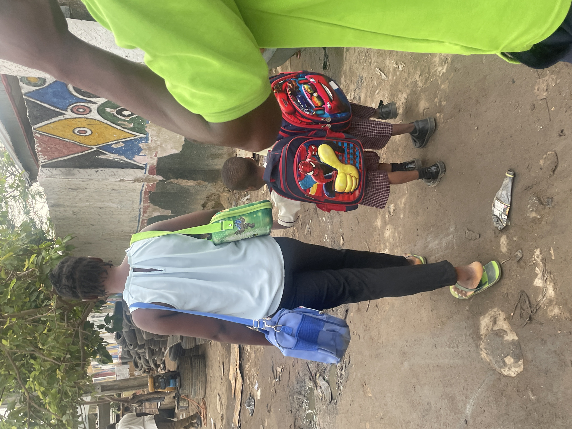 Two children with backpacks, one blue Spiderman themed and one red with other characters, stand on a dirt path. A woman in a light blue shirt, black pants, and green flip-flops walks alongside them, carrying a light green and blue purse. An adult in a neon green shirt stands nearby. The background reveals a weathered building with colorful geometric designs and a pile of tires. The location is Abuja, Nigeria. The scene is dusty and cluttered.