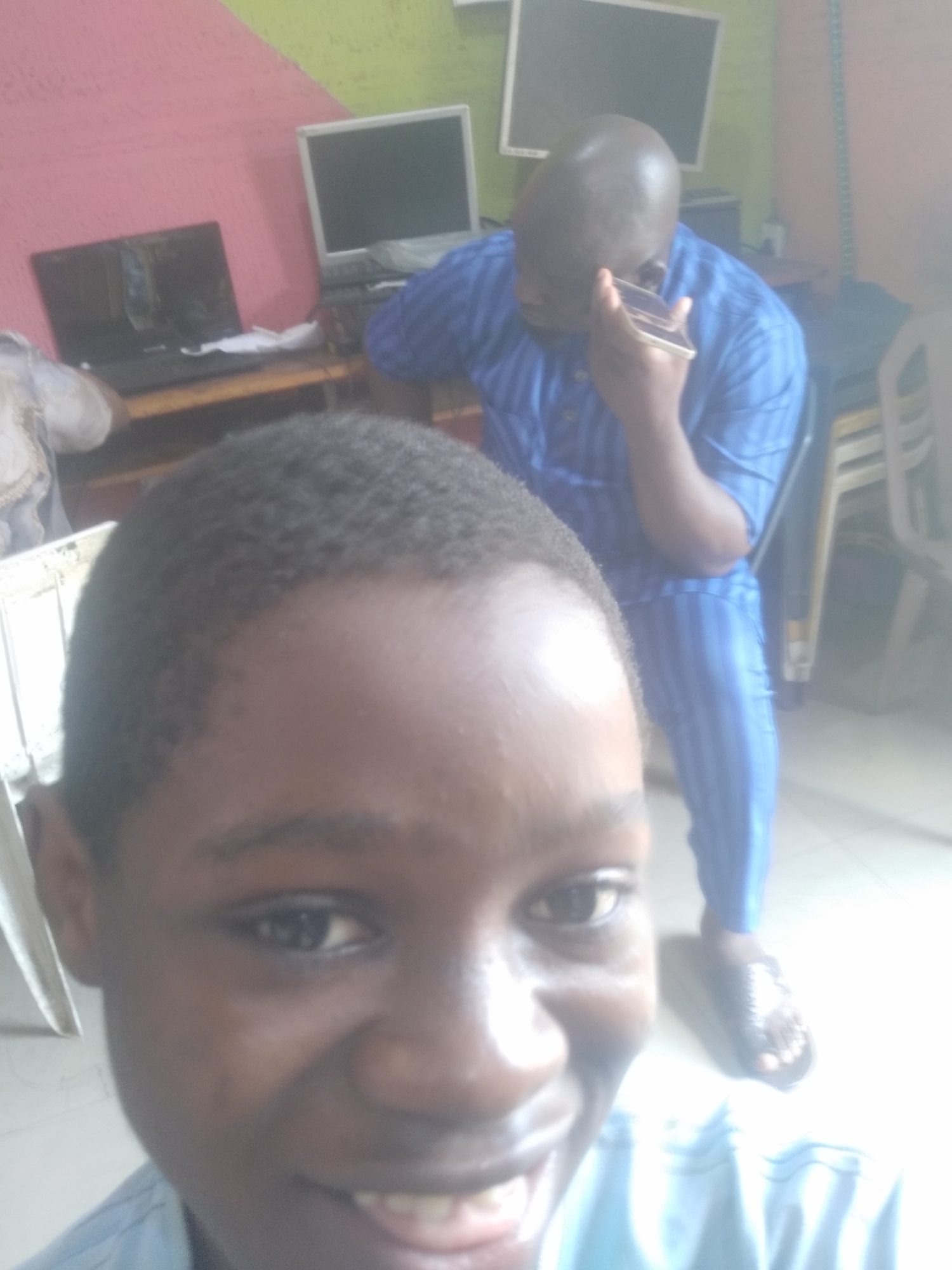 The image shows a young person taking a selfie. Behind them stands an older, bald man dressed in a blue striped shirt and pants. He is holding a phone to his ear. In the background are several computers and some stacked chairs. The location is Unknown, Unknown.