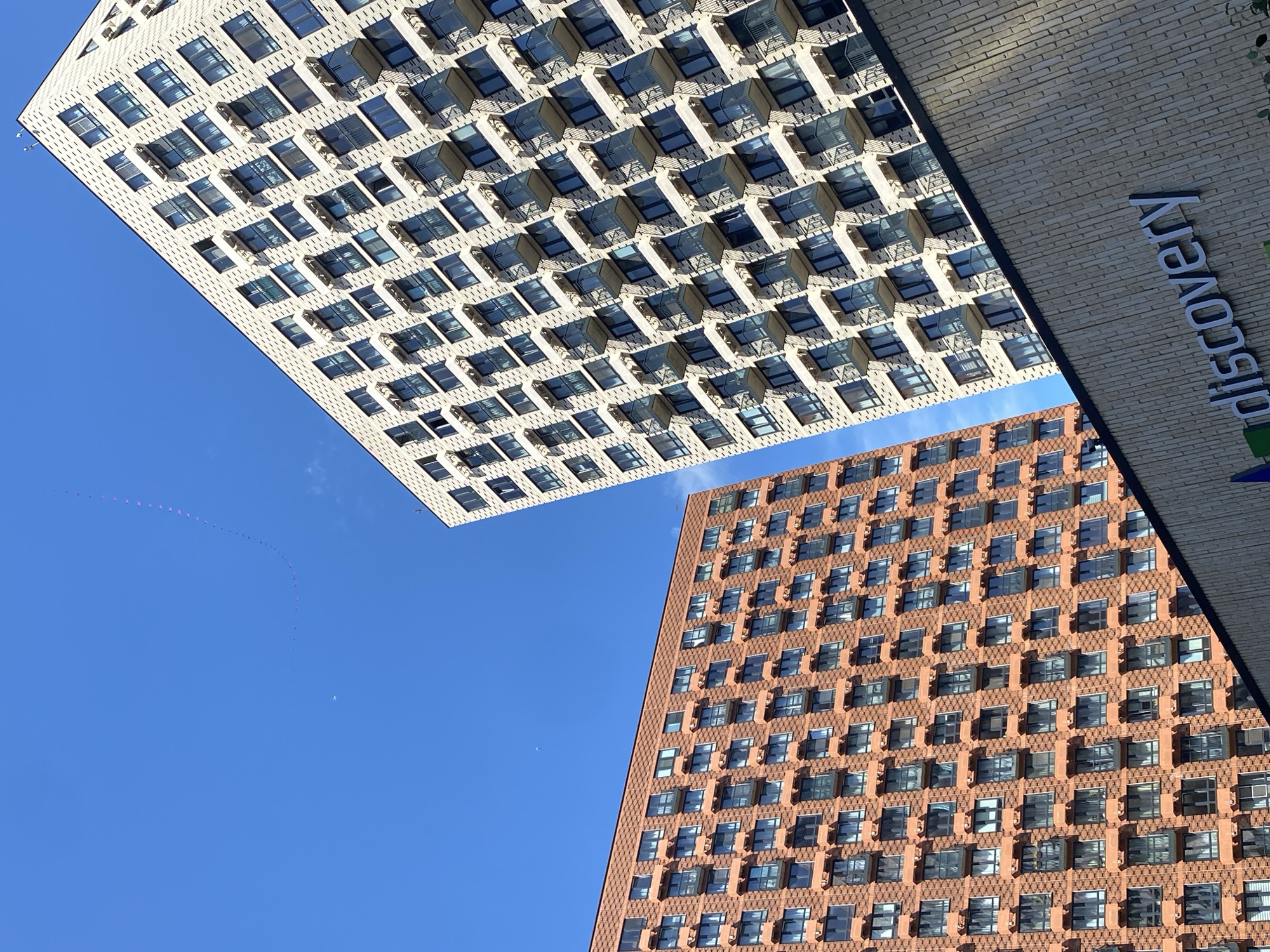 The image depicts a sunny day in a modern urban environment, likely Moscow, Russia, as suggested by the architecture and the presence of the "discovery" logo on a building facade. Two tall residential buildings dominate the frame, angled against a clear blue sky. The building on the left is clad in reddish-orange bricks and features numerous windows and balconies. The building on the right has a lighter, cream-colored facade with a patterned texture, also replete with windows and balconies. A lower, modern building with a textured facade, bearing the prominent "discovery" logo in silver lettering, occupies the foreground, partially obscuring the base of the right-hand building. In the upper portion of the sky, a string of small, colorful flags or pennants is visible, likely attached to a kite or drone, trailing in a curve against the blue. A small, faint white object, possibly an airplane, is also discernible in the sky. There are no people or vehicles clearly visible in the image.