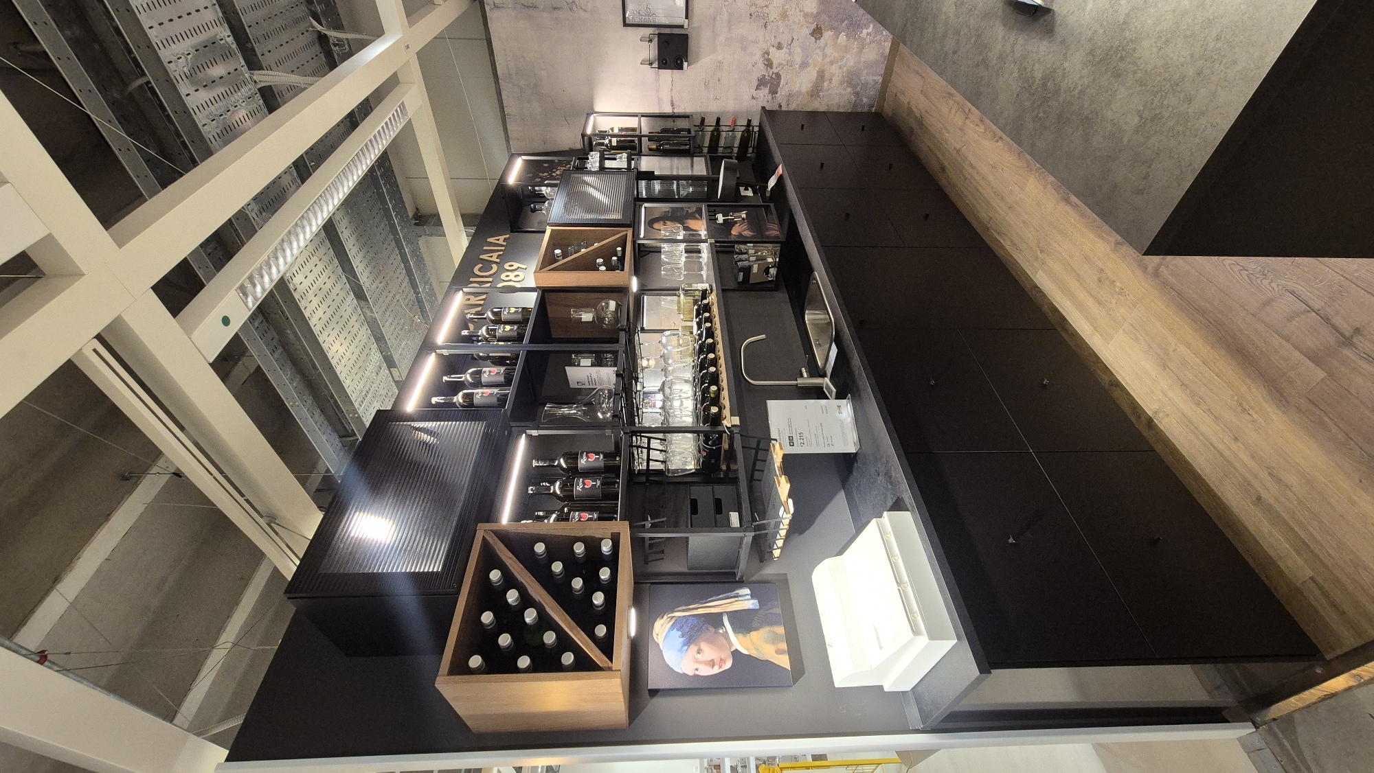 The image depicts a modern display kitchen or bar area. The scene is dominated by dark cabinetry and shelving units, featuring a collection of wine bottles, decanters, and glassware. A painting of "Girl with a Pearl Earring" is prominently displayed on the left wall, next to a white cash register or dispenser. The countertop is black, with a stainless steel sink and faucet. Above the counter, illuminated shelves showcase more bottles and decorative items. The ceiling structure is visible, with exposed conduits and lighting. The flooring is a light-colored wood laminate. A price tag is attached to the shelving unit near the sink. The overall aesthetic suggests a retail or showroom environment, possibly in Baronissi, Italy, given the context. There are no people visible in the image.