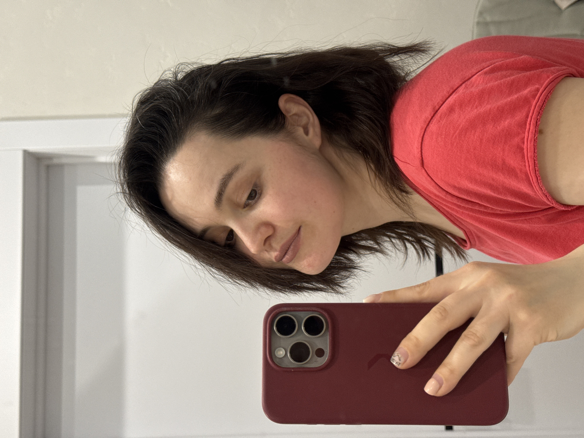 A woman with dark hair wearing a red t-shirt is holding a red phone with a camera in her right hand and taking a selfie. She has fair skin and is looking down at the phone. The background is a neutral wall and a white door frame. There is no indication of Izhevsk, Russia, or any other specific location.