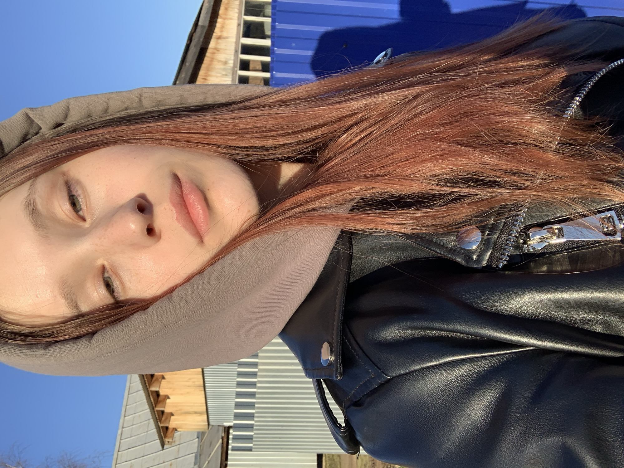 This close-up, diagonally oriented outdoor portrait features a young person, likely female, captured in bright daylight in Фоки, Russia. The subject's face, primarily the left side from forehead to chin, is visible in the lower-left portion of the frame. She has fair skin with visible freckles on her nose and cheek, and light-colored eyes, possibly green or grey-green. Her long, reddish-brown hair cascades across her shoulder and chest.
She is dressed in a grey hooded top, with the hood framing part of her face, and a black leather jacket adorned with prominent silver zippers and snaps. The strong sunlight creates clear highlights on her skin and hair, and on the metallic details of her jacket. Her expression is neutral and calm, with her gaze directed slightly away from the camera.
The background shows a clear, bright blue sky in the upper left, indicating a sunny day. To the upper right, a distinct blue corrugated metal structure, possibly a wall or container, is visible, with a dark shadow cast upon it. Further into the background, wooden planks and other light-colored corrugated metal sheets suggest parts of buildings or fences. The bright lighting and clear shadows indicate it is daytime, likely mid-day or early afternoon. No visible text is present in the image.