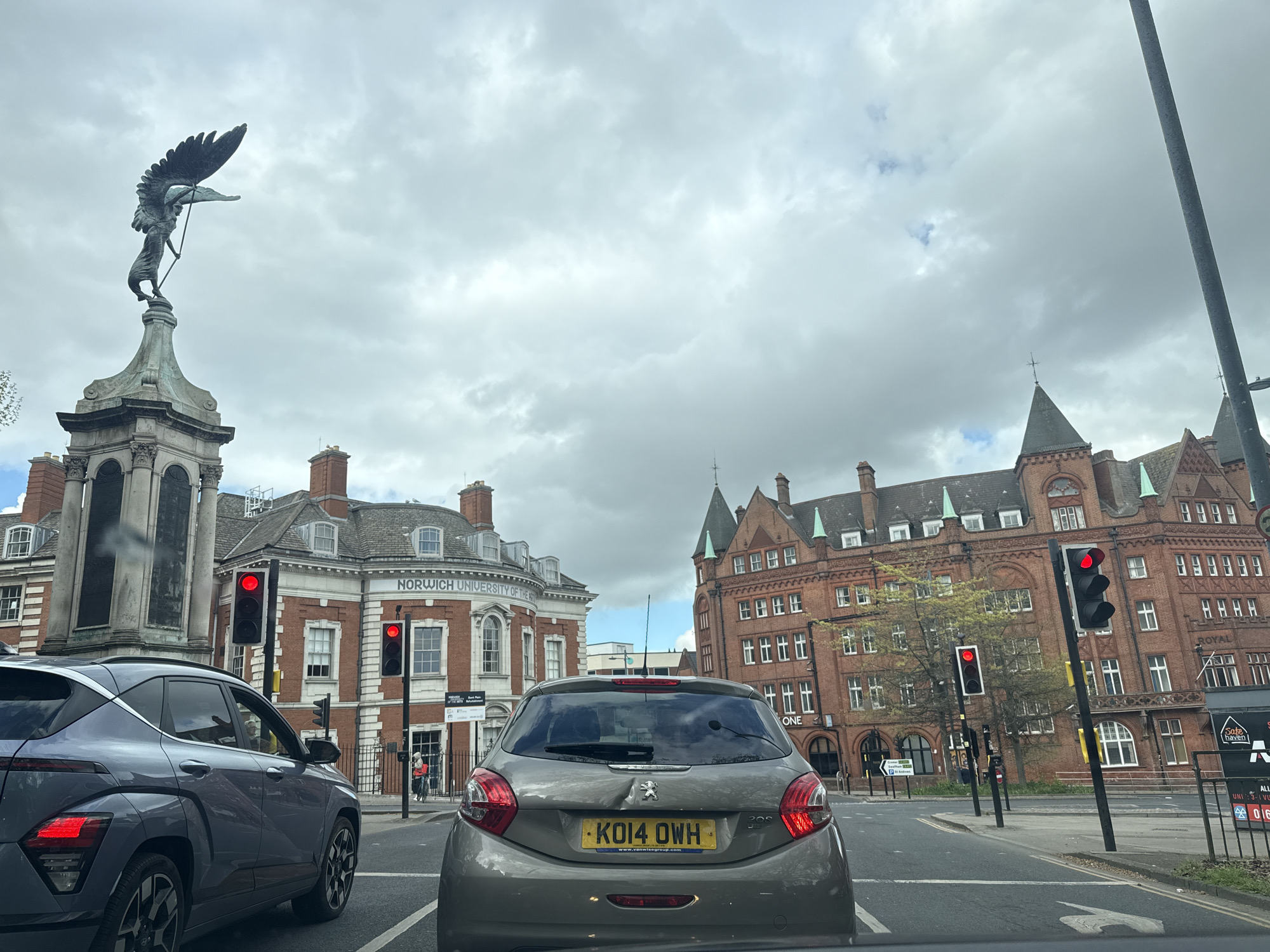 The image shows a street scene in Norwich, United Kingdom, captured from inside a vehicle. In the foreground are two cars stopped at a traffic light. The car in the immediate foreground is a Peugeot 208 with the license plate "KO14 OWH". To the left is a grey Hyundai. All traffic lights in view are red. In the background are several prominent buildings, identifying the location as near Norwich University of the Arts (building on the left) and the Royal Arcade (building on the right). A war memorial with a winged statue sits to the left. The sky is overcast with grey clouds. There is a pedestrian visible on the left side of the frame near the Norwich University of the Arts building. The road appears to be dry. Signage indicating businesses like "Sake Haven" can be seen in the background.