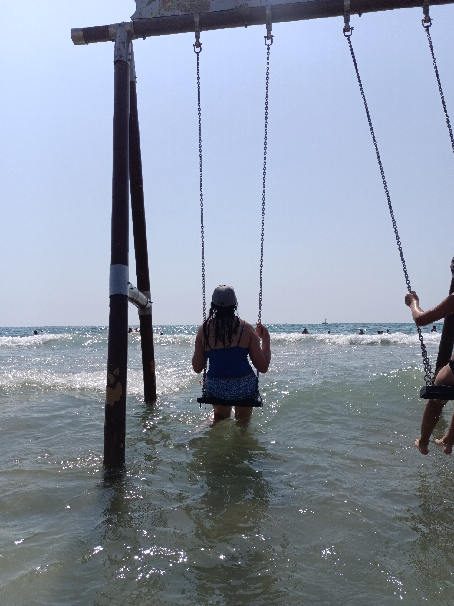 In La Playa, Spain, two individuals are seen on swings partially submerged in the ocean. The person on the left, wearing a blue top and patterned shorts, is seated on a swing. They are facing away from the camera, with their back to the viewer. A grey baseball cap is worn on their head. The swing's chains are attached to a metal frame structure. The person on the right is also seated on a swing, with their legs visible as they are partially submerged in the water.
The ocean features moderate waves, with some whitecaps indicating movement. Sunlight reflects off the water's surface, creating bright glints. In the background, other people are visible in the water, some swimming and one individual on a small boat or raft. The sky is clear and blue, indicating a sunny day. The presence of a swing set at the ocean's edge suggests a recreational area.