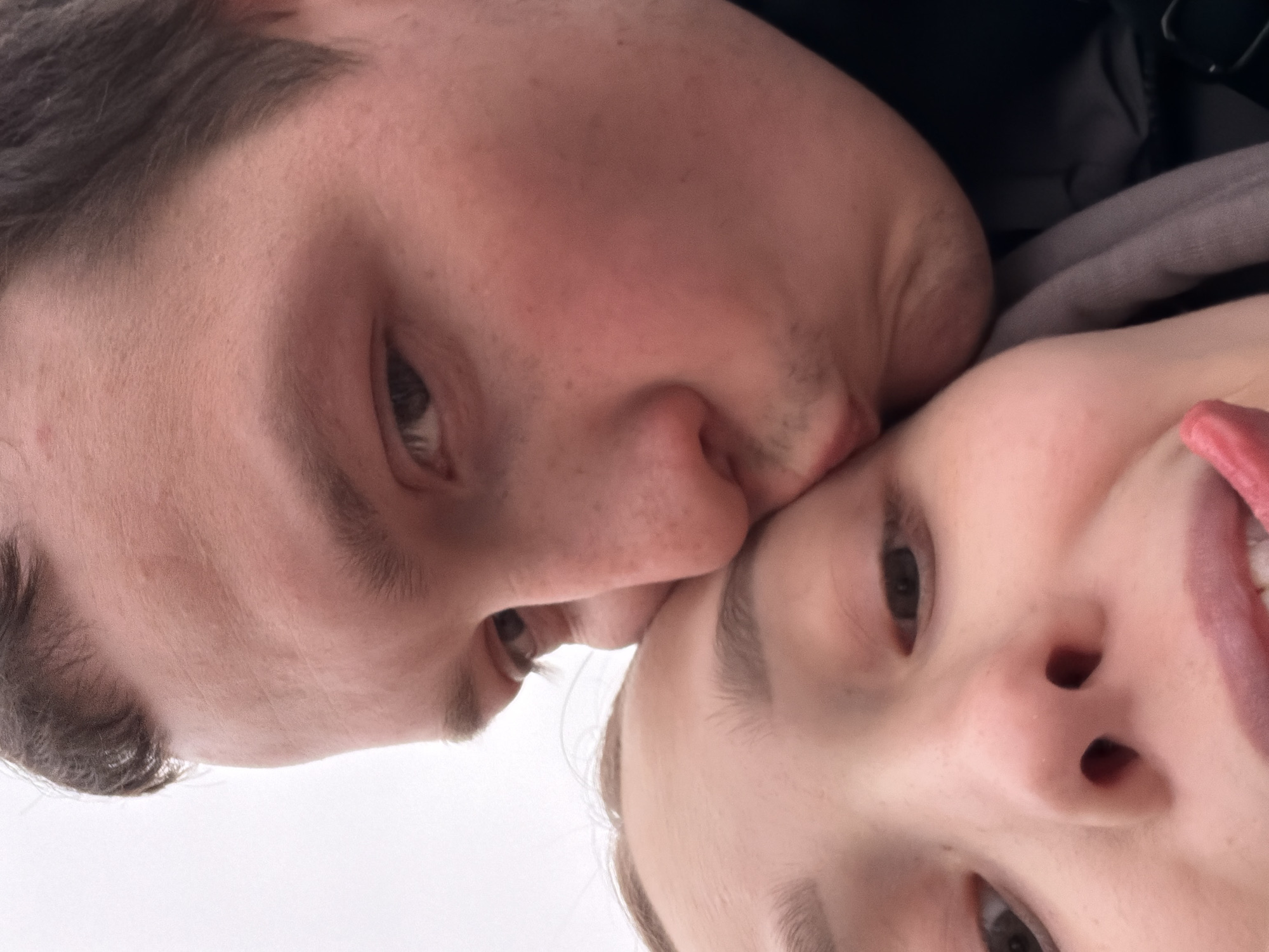 A close-up, off-angle shot depicts two individuals, presumably in a romantic relationship, with the male figure kissing the female figure's forehead. The male has short, dark hair, light skin, and visible stubble. The female has fair skin, dark eyes, and a partly visible tongue. The background is indistinct, but potentially out-of-doors under overcast conditions. The location is unknown, and the specified city of Chaikovsky, Russia, cannot be confirmed from the visual information provided.