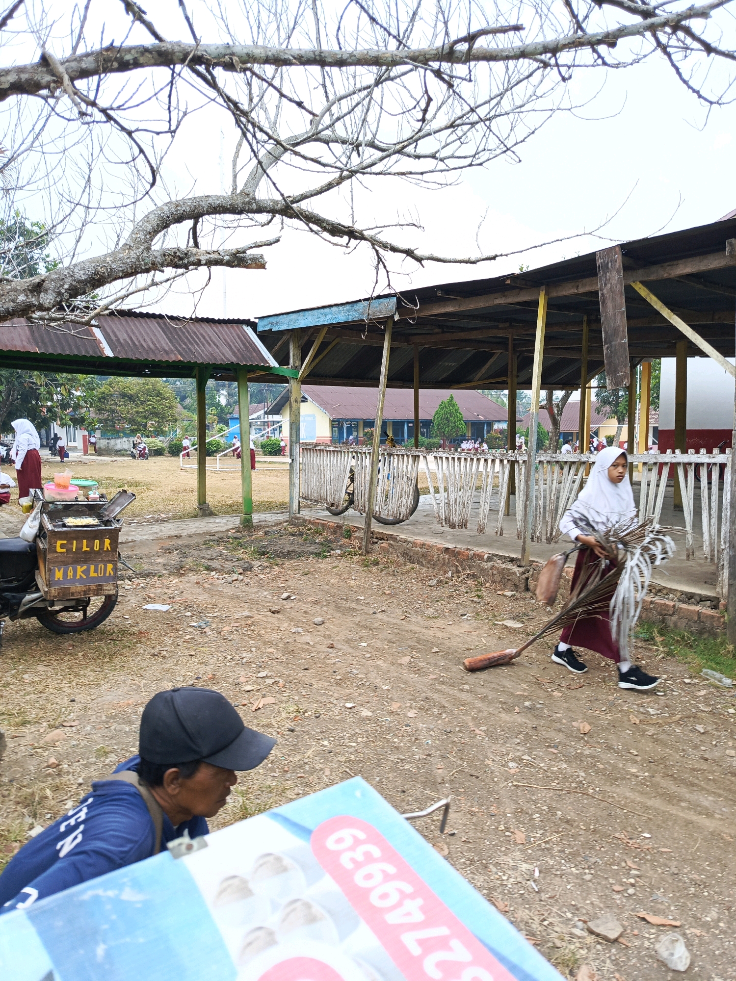 The image depicts an outdoor scene likely on the grounds of a school in Megang Sakti II, Indonesia. In the foreground, a person wearing a blue hoodie and a black baseball cap is partially visible, facing away from the camera. To the left, a motorcycle-mounted food cart with the text "CILOR MAKLOR" is parked. In the mid-ground, a young girl in a white hijab and maroon school uniform is sweeping the dirt ground with a broom made of palm fronds. She is carrying a bundle of dried palm leaves. Behind her, a covered outdoor structure with a corrugated metal roof and wooden support beams is visible. Several other students in similar uniforms are seen in the background, some walking and others near buildings that appear to be classrooms. The environment is dry and dusty with sparse vegetation and a partly cloudy sky.