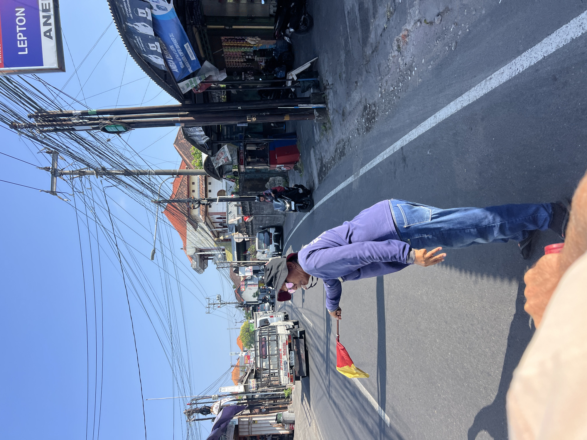 A person in a purple long-sleeved shirt and blue jeans, wearing a black hat and pink sunglasses, is holding a red and yellow flag in their right hand, directing traffic on a paved road. The person is positioned on the right side of the road. In the background, there is a line of vehicles, including a white truck with license plate "DK 0367 BY", a white van, and several motorcycles. Shops and buildings line the street, with power lines and utility poles overhead. The sky is clear and blue. The scene appears to be a street in Denpasar, Indonesia, possibly near a busy intersection or construction zone where traffic control is needed. The overall atmosphere is one of a typical day in a bustling urban environment.