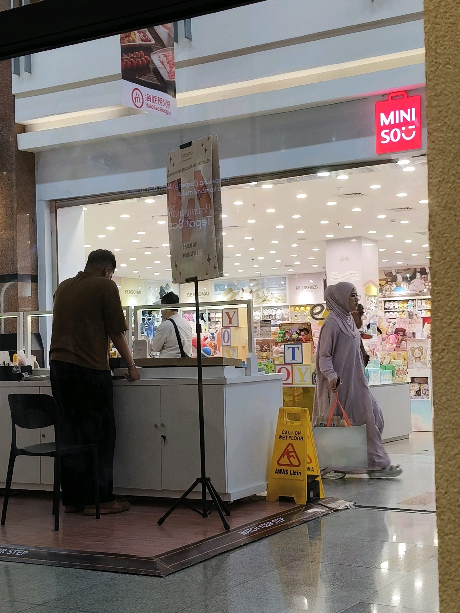 In Surabaya, Indonesia, inside a shopping mall, a man in a brown shirt and black pants stands behind a counter. To his right, a woman wearing a light purple hijab and dress walks by, carrying a light gray tote bag with orange straps. She is walking past a yellow wet floor sign. Behind the counter, shelves are filled with various merchandise, and a red neon sign with white lettering that reads "MINI SO" is visible above the entrance to a store. A banner advertising "Haidilao Huoguo" hangs on the wall above the store. The floor is tiled and reflects the overhead lighting.