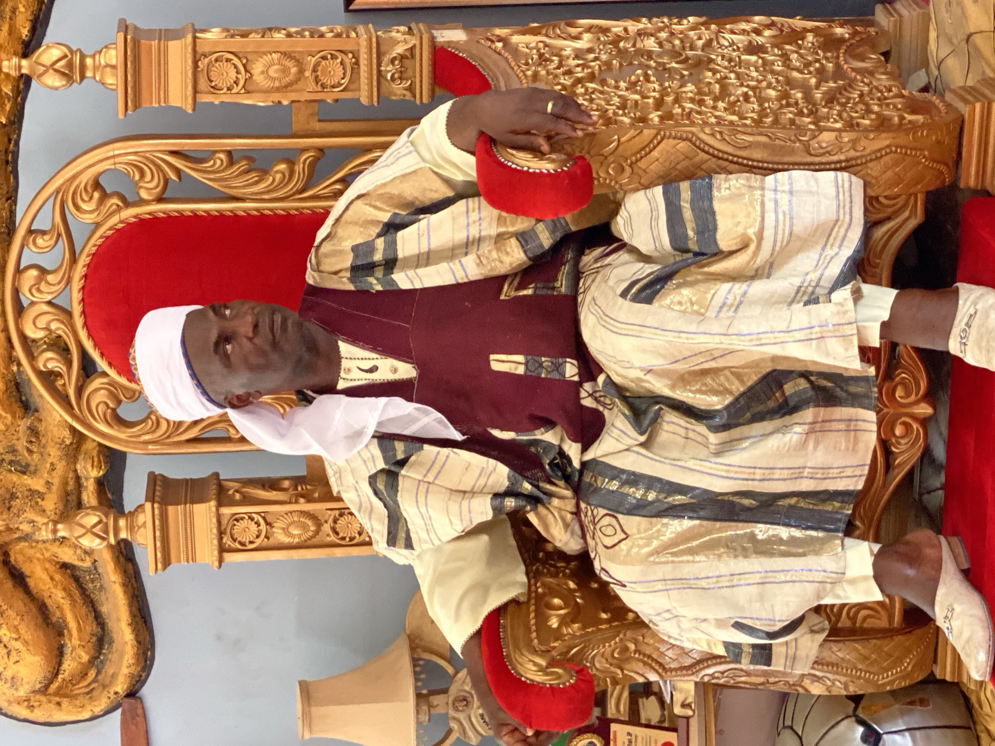 A man wearing a traditional outfit and headdress sits on an ornate, gilded throne with red upholstery. He holds a small red object in his hand. The throne's backdrop is adorned with detailed carvings and the room seems to be in a place of authority, possibly Sabon Tasha, Nigeria.