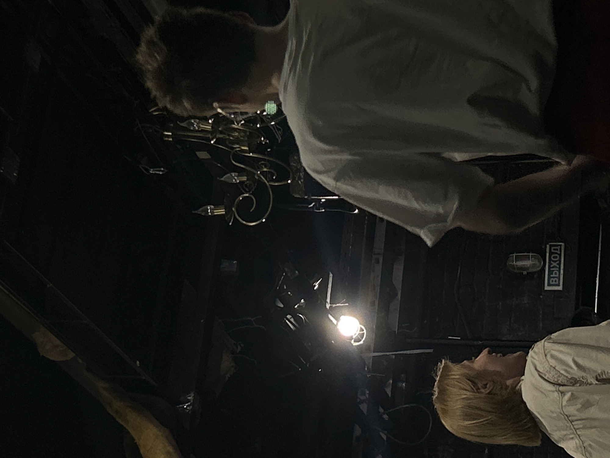 In an interior space with dark, exposed ceiling beams and brick walls, two individuals are visible. A woman with blonde, shoulder-length hair is on the left, looking to her right with an open-mouthed smile. She is wearing a light-colored jacket. To her right and slightly in front of her, a person with short, brown hair, seen from the back, is wearing a white t-shirt. Behind them, a chandelier with multiple lights hangs from the ceiling. A bright spotlight illuminates a section of the ceiling and a metal structure. On the brick wall to the right of the woman, a light fixture is mounted above a sign that reads "ВЫХОД" (Exit in Russian). The scene appears to be dimly lit, suggesting an evening or indoor event.