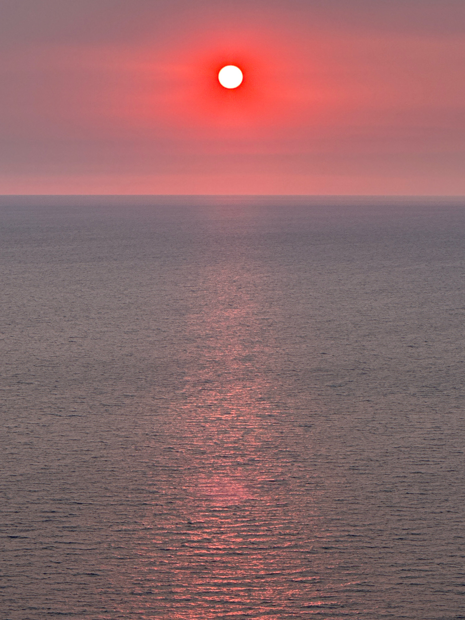 A vertical, medium shot captures a hazy red sun, partially obscured by atmospheric conditions, positioned in the upper center of the frame. The sun illuminates a vast expanse of calm, grey-purple water, which fills the lower two-thirds of the image. A shimmering reflection of the sun stretches across the water's surface, creating a path of pinkish light. The horizon line is sharp and distinct, separating the muted sky from the tranquil sea. The location is identified as Glavatičići, Montenegro. There are no people, animals, or man-made structures visible in the image.