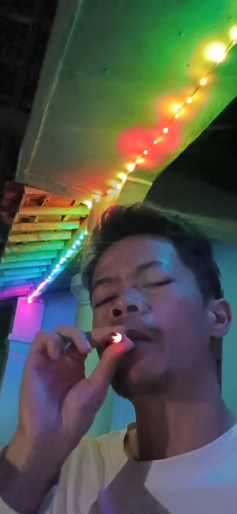 A young man is taking a selfie with a cigarette, holding it up to his lips. The scene appears to be outdoors, with colorful LED lights strung along a ceiling or overhang. The lights cast a vibrant glow of green, yellow, red, and blue on the man's face and surroundings. In the background, wooden planks are visible, possibly part of a building or structure. The man is wearing a white t-shirt. The image quality is somewhat grainy, suggesting it may have been taken in low light conditions. The location is identified as Wonokerso, Indonesia.